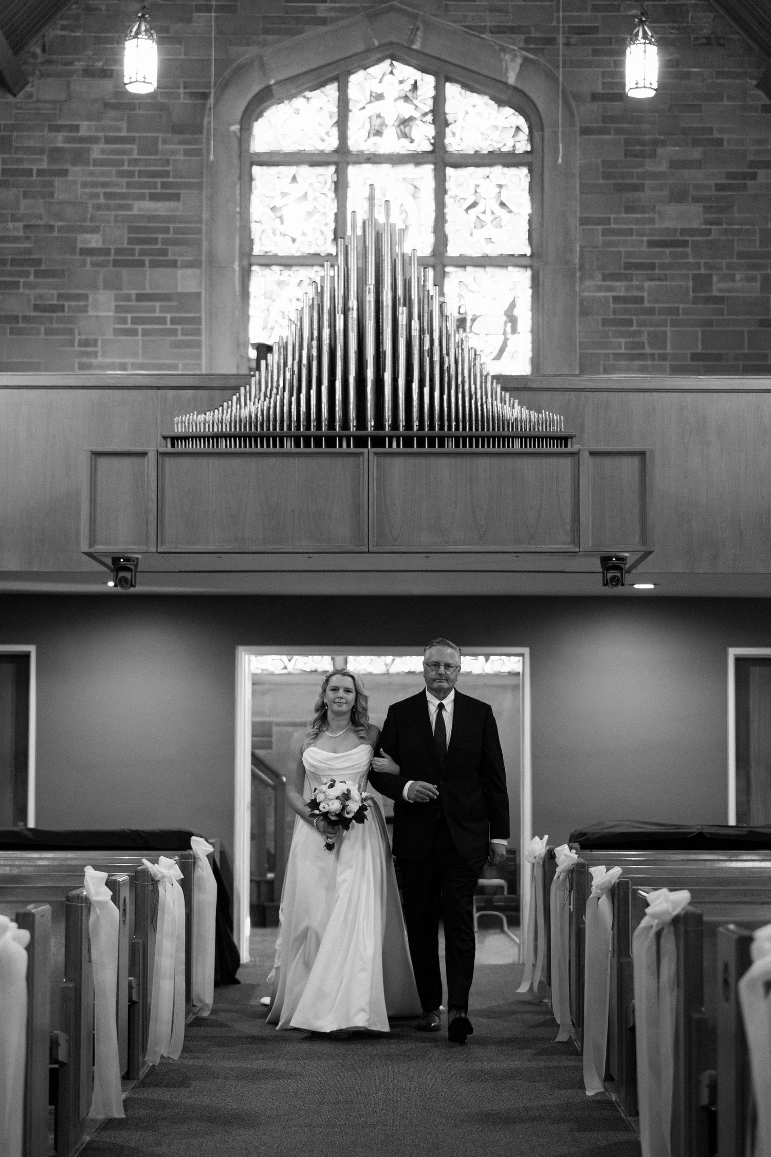 Black and white photo of a bride walking down the church aisle with her father past wooden pews toward the altar.