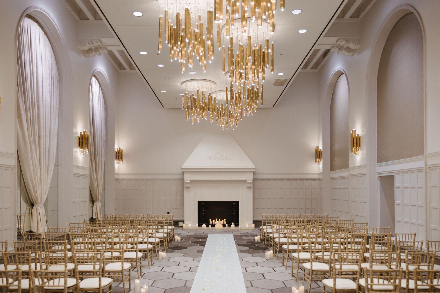Elegant wedding ceremony venue with gold chandeliers, chiavari chairs, white aisle runner, and a fireplace in a grand ballroom.