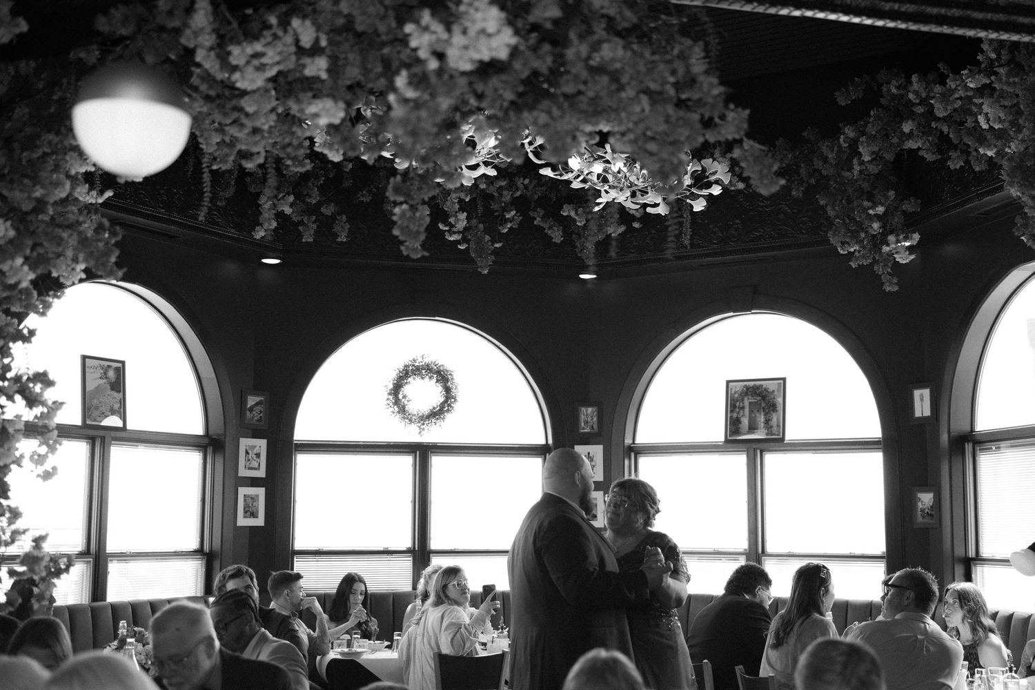 Black and white wedding reception photo with couple dancing under floral ceiling in elegant arched window venue.