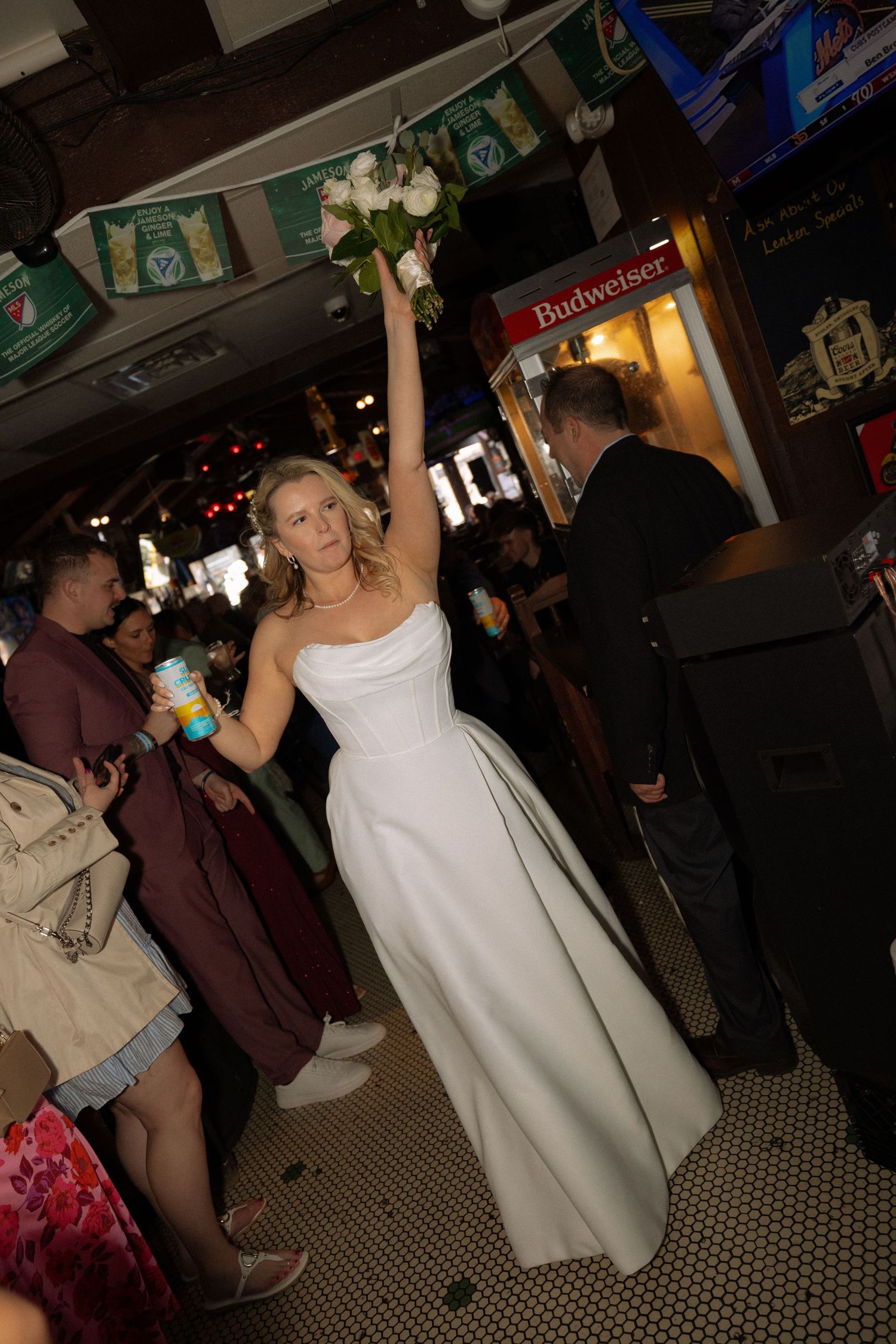 Bride in white gown raises bouquet triumphantly inside a bar with Budweiser signage during wedding celebration.