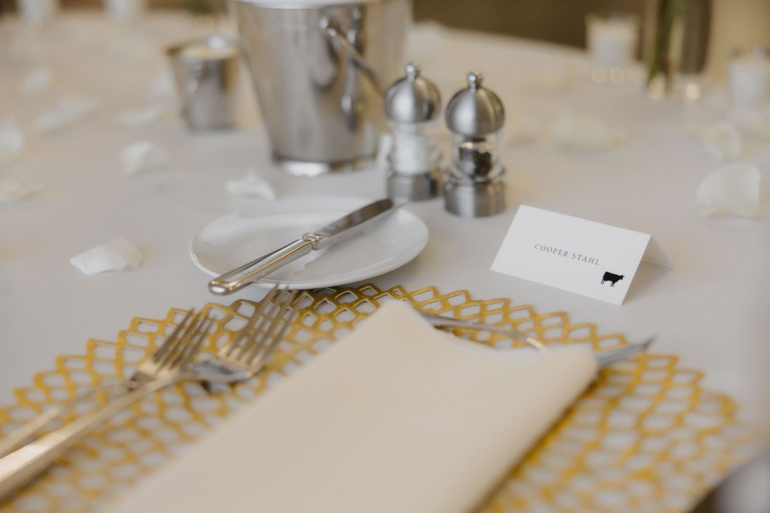 Elegant wedding table setting with gold geometric placemat, silverware, white plate, name card, and silver salt and pepper shakers.