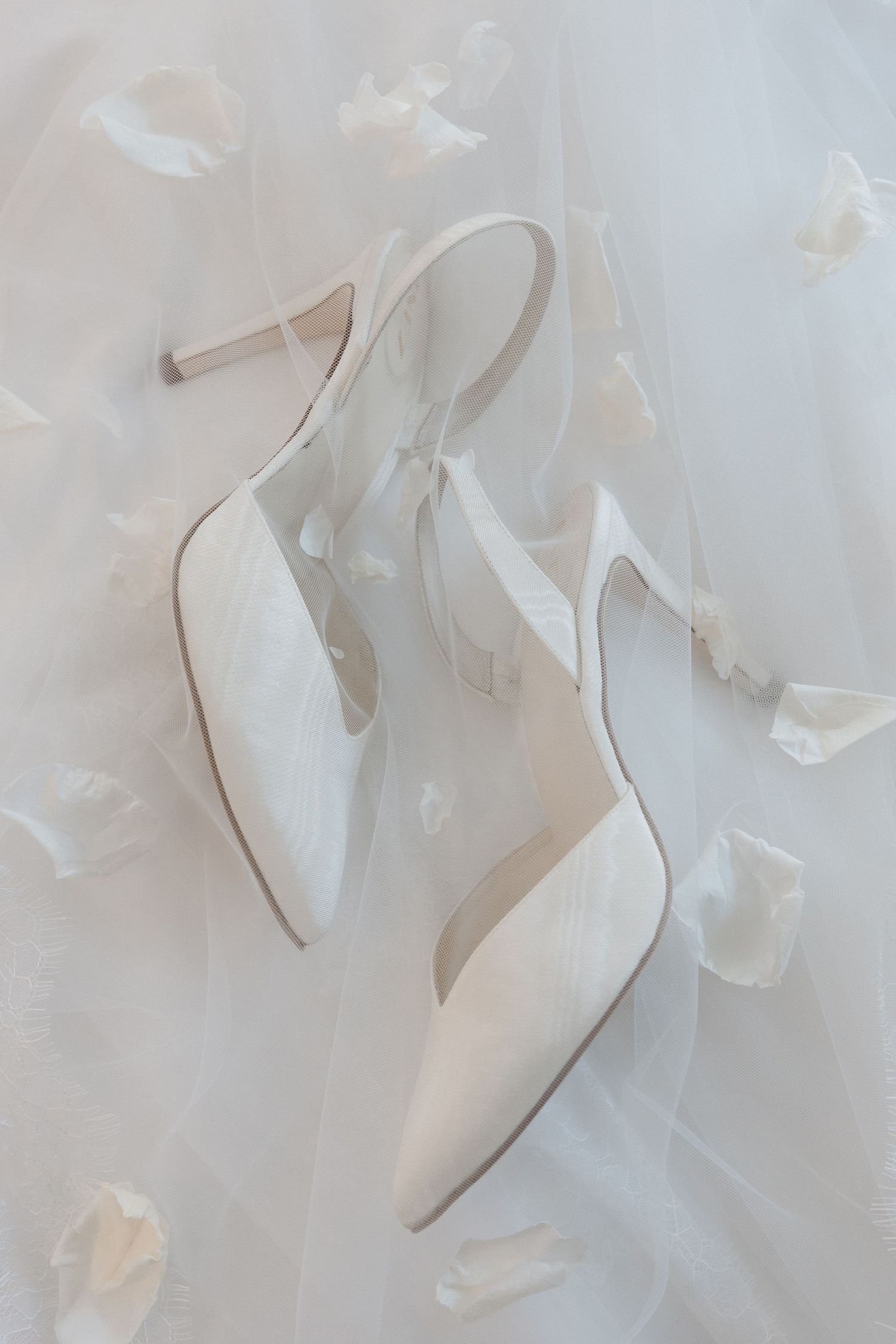 Elegant white satin slingback heels with ribbon ties resting on a bridal veil surrounded by soft flower petals.