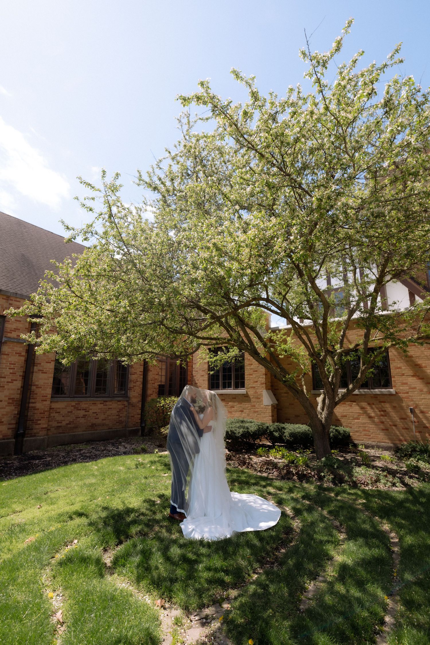 Bride and groom kissing under a blooming tree in a lush garden beside a brick building on a sunny wedding day.