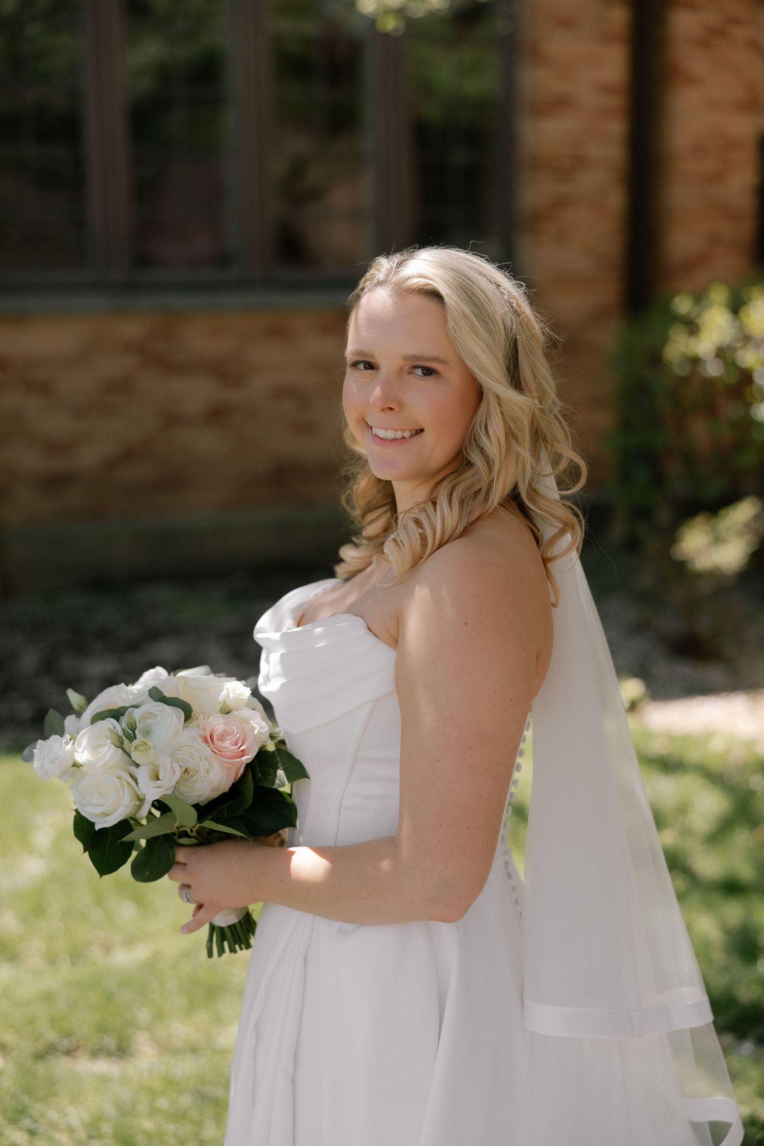 Smiling bride in white strapless gown holding pink and white bouquet with veil outdoors.
