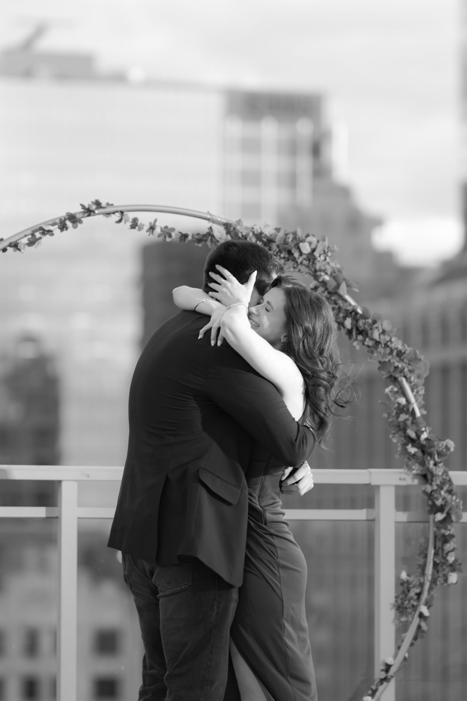 Black and white photo of couple embracing passionately under a floral arch on a city rooftop.