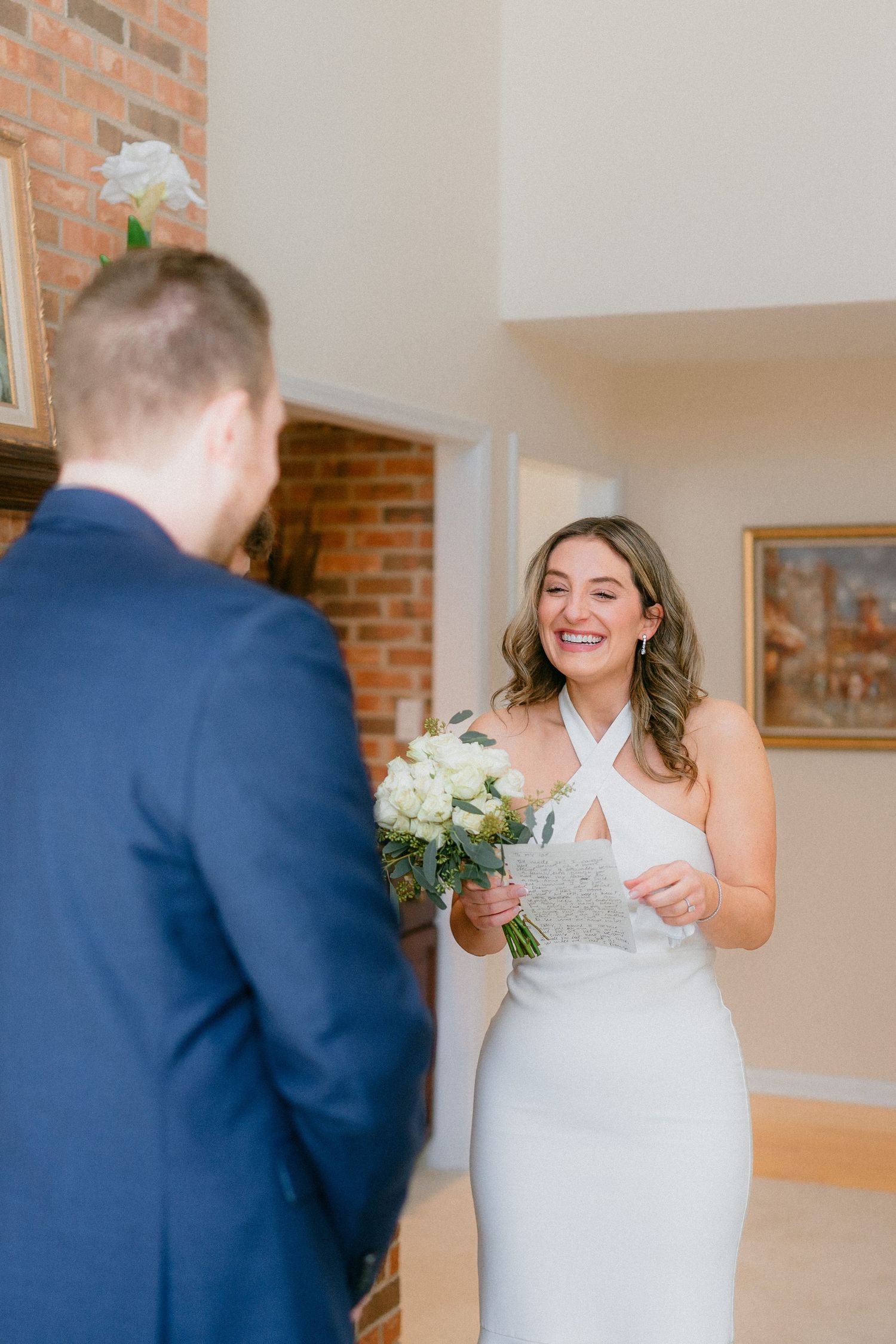 Radiant bride in white halter gown smiling at groom while holding bouquet and vows.