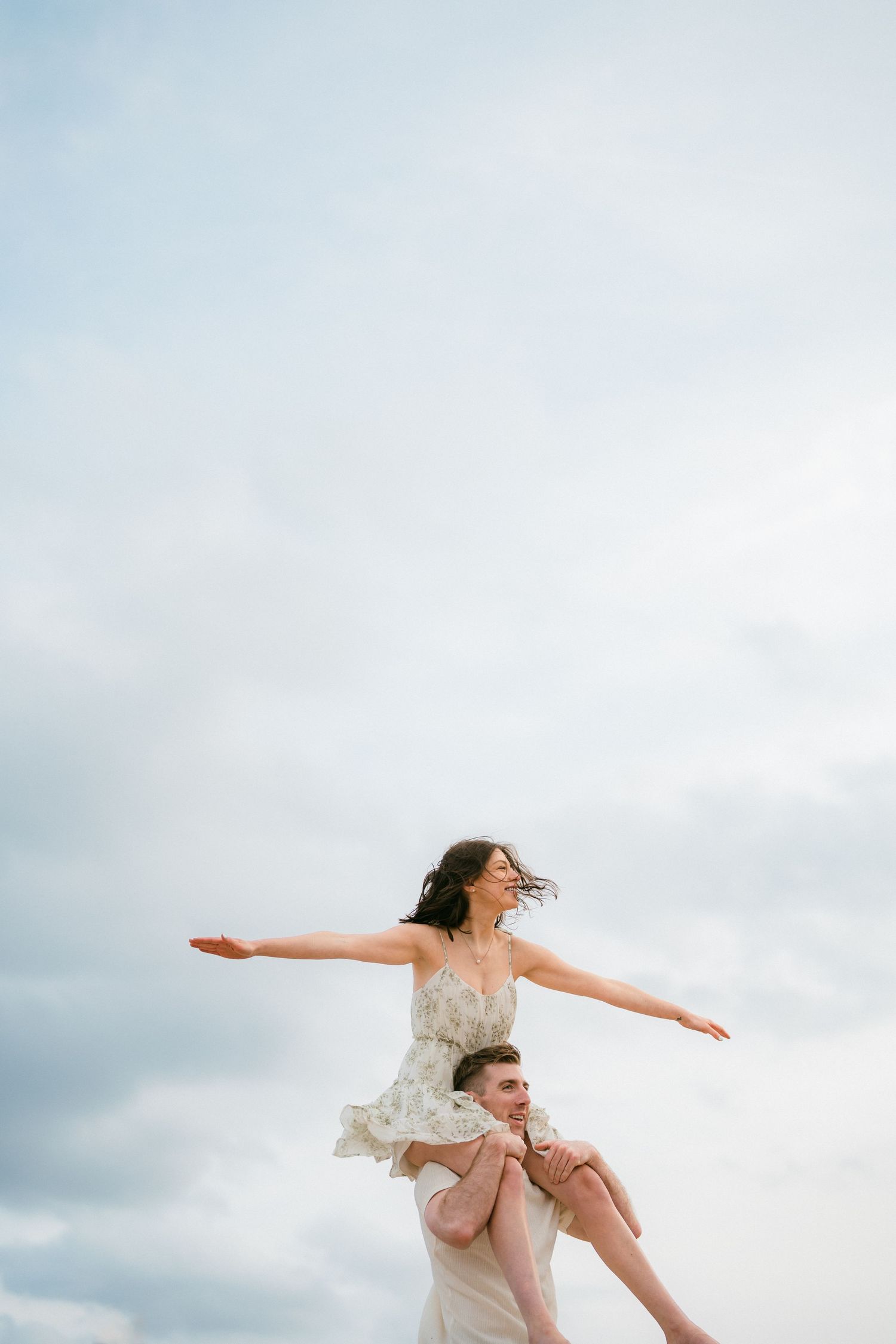 Woman sitting on man's shoulders with arms spread wide against a dramatic cloudy sky.