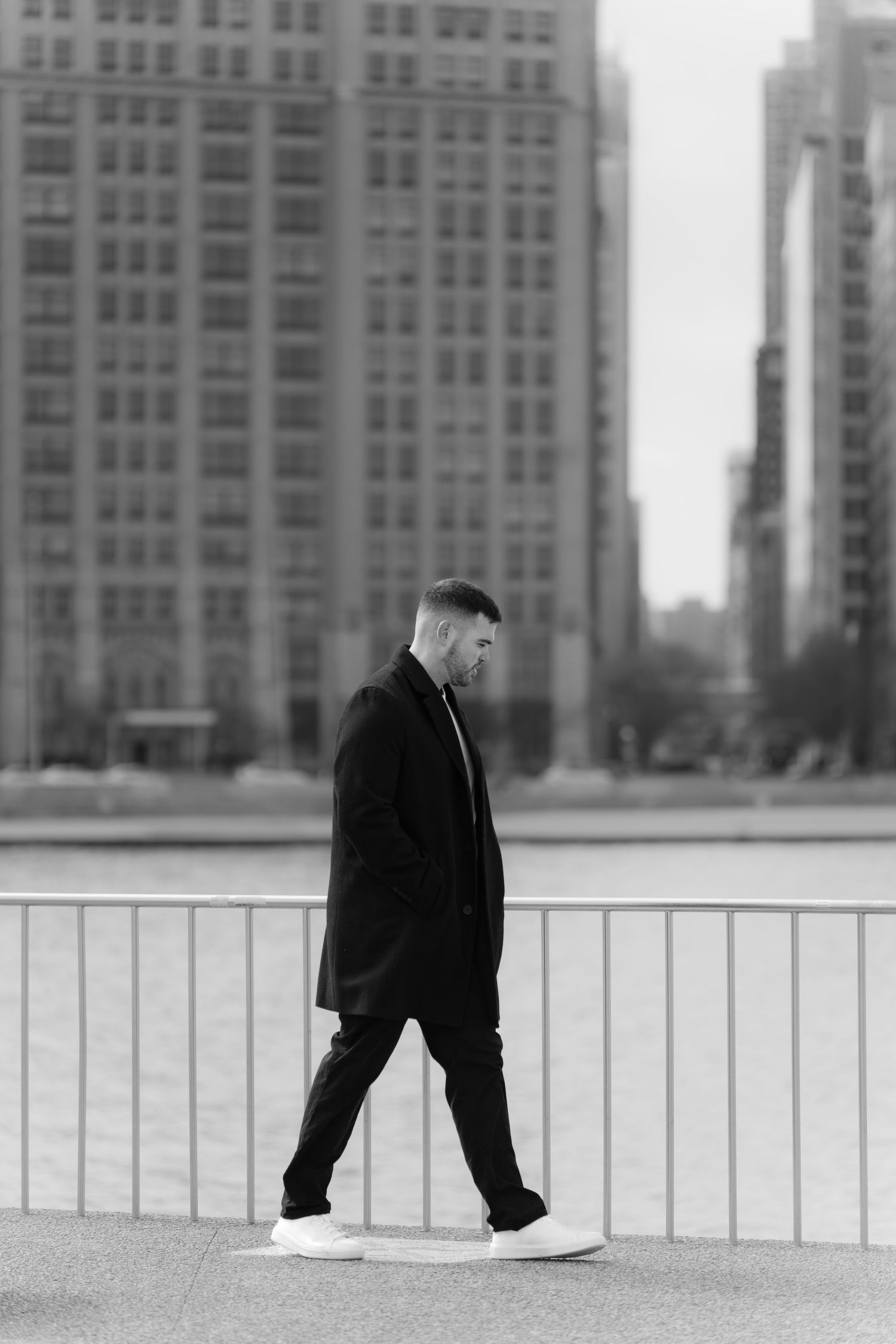 Stylish man in black coat walking along urban waterfront railing in black and white street photography.