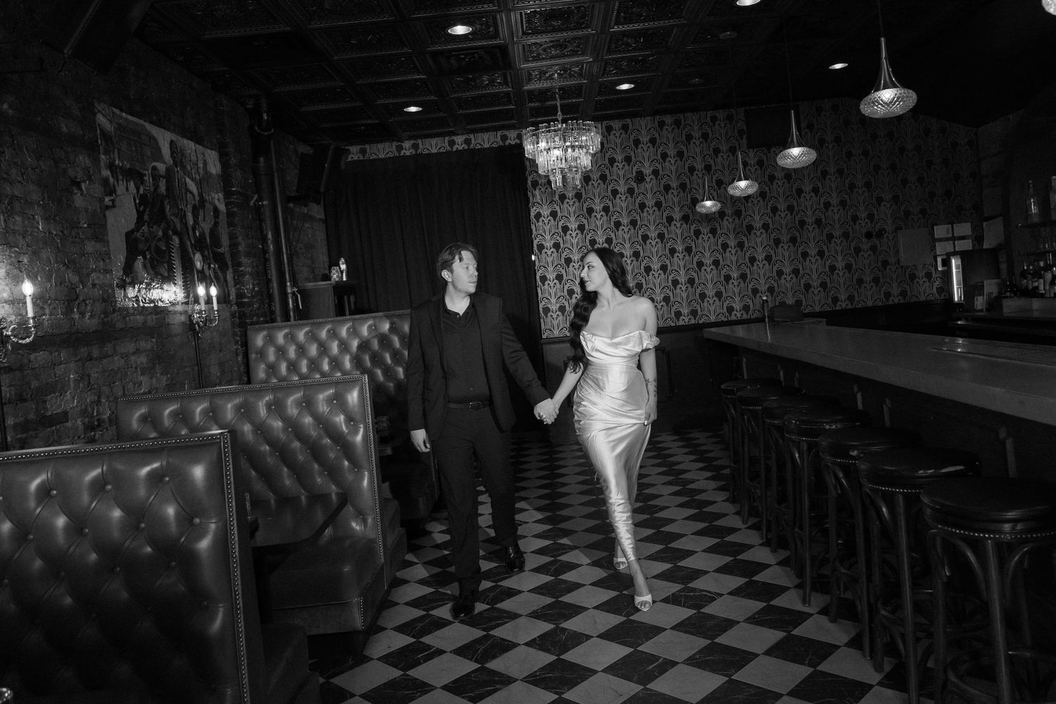 Couple holding hands walking through elegant black and white checkered floor bar with chandelier lighting.