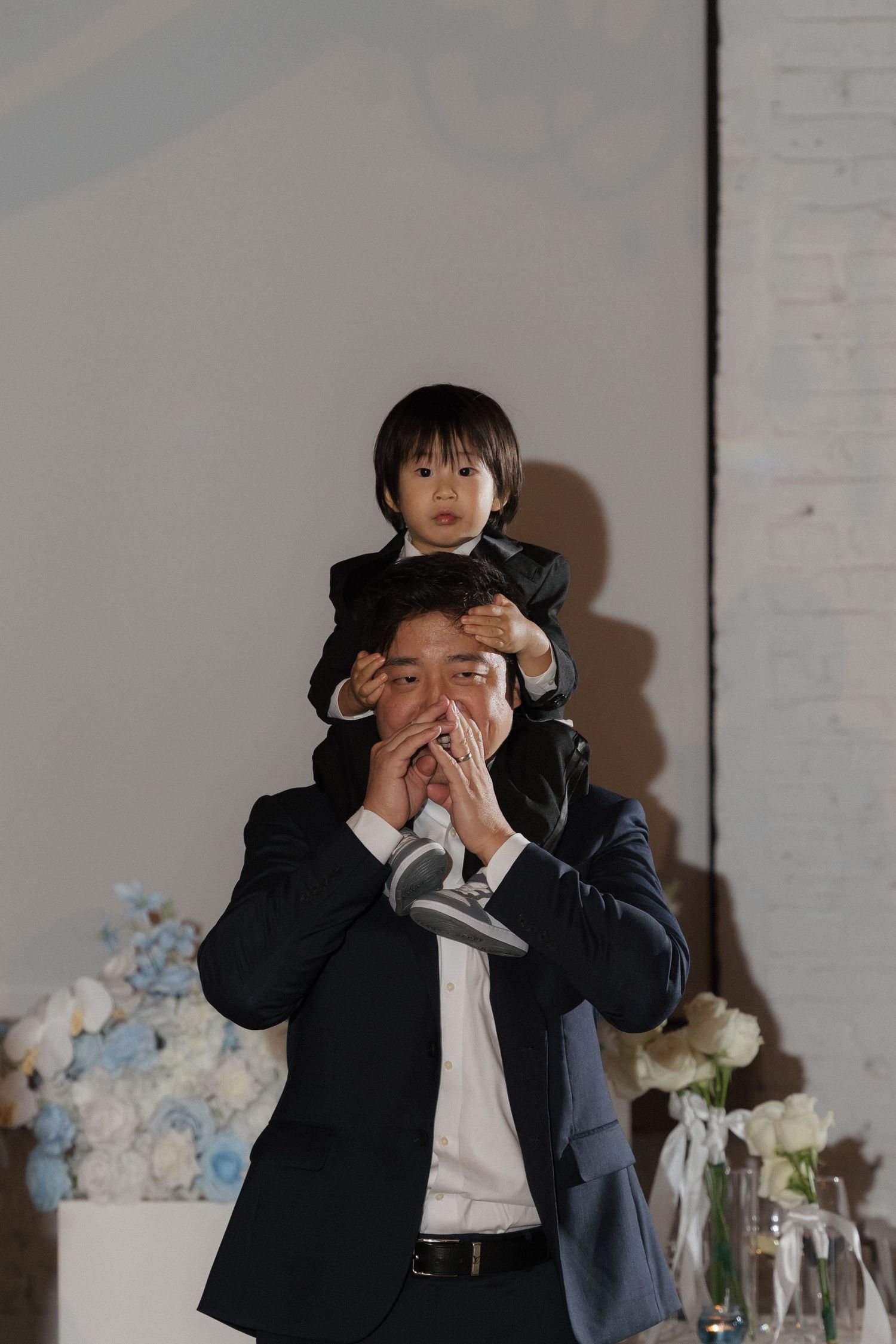 Man in navy suit holding young child on shoulders at wedding reception with floral backdrop.