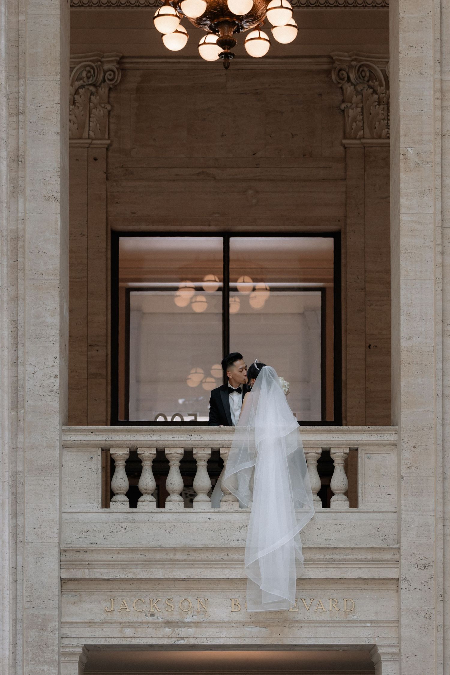 Bride and groom share romantic kiss on ornate balcony of historic wedding venue.