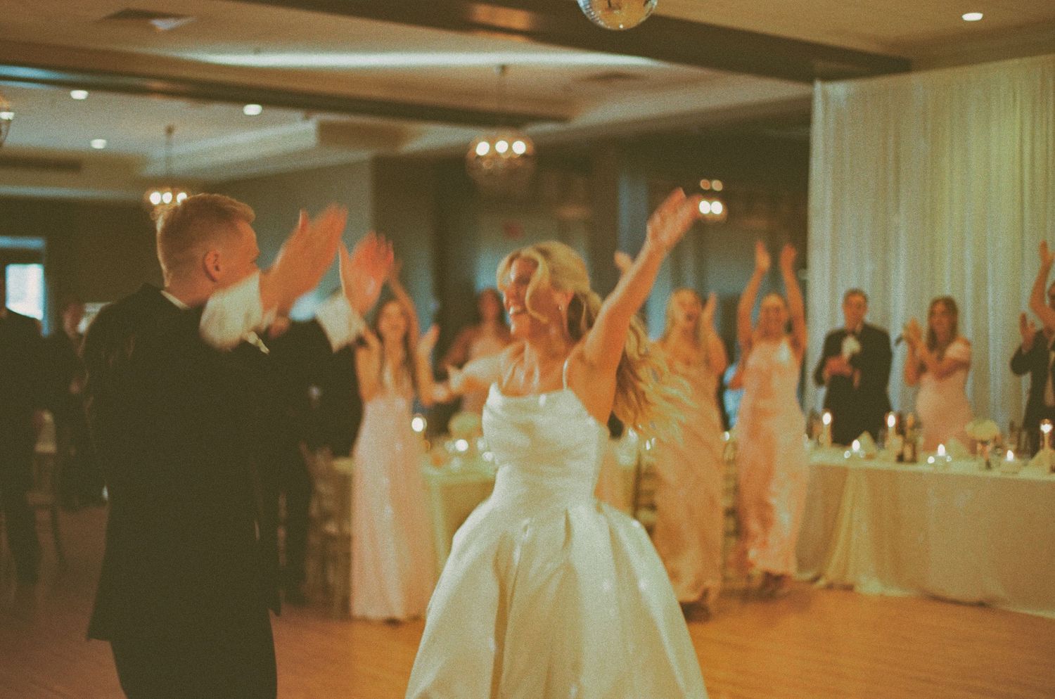 Bride and groom share first dance at wedding reception with guests clapping in elegant ballroom.