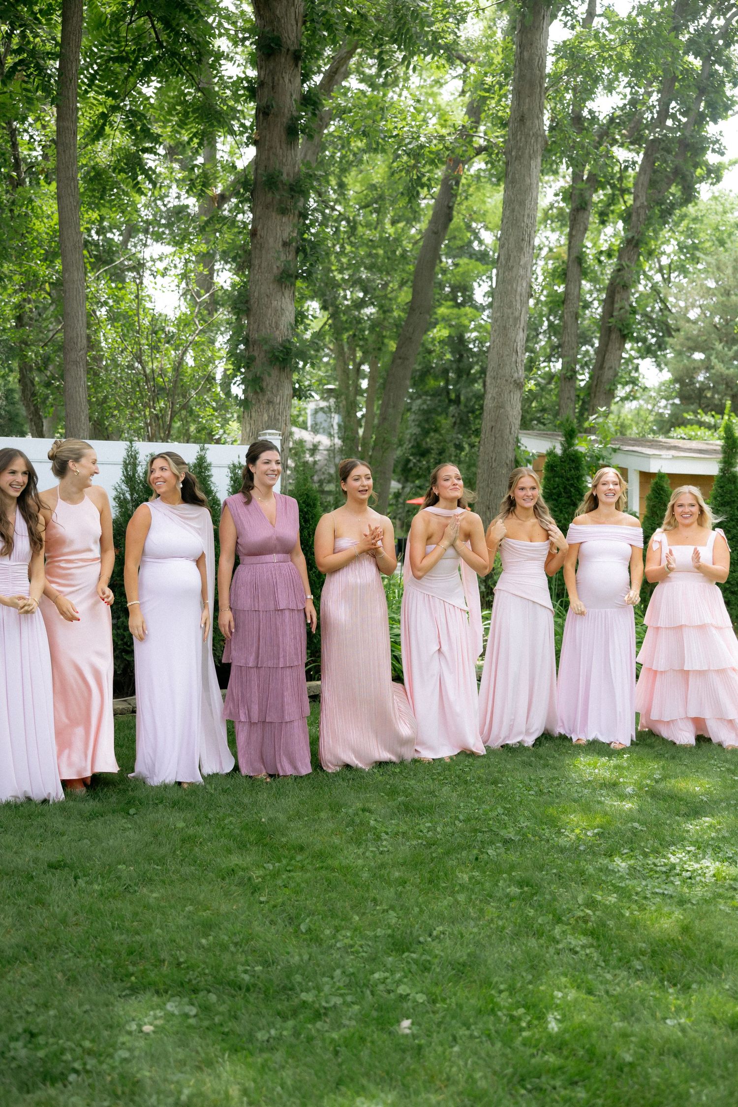 Eight bridesmaids in pink dresses standing on green lawn with trees in background at outdoor wedding.
