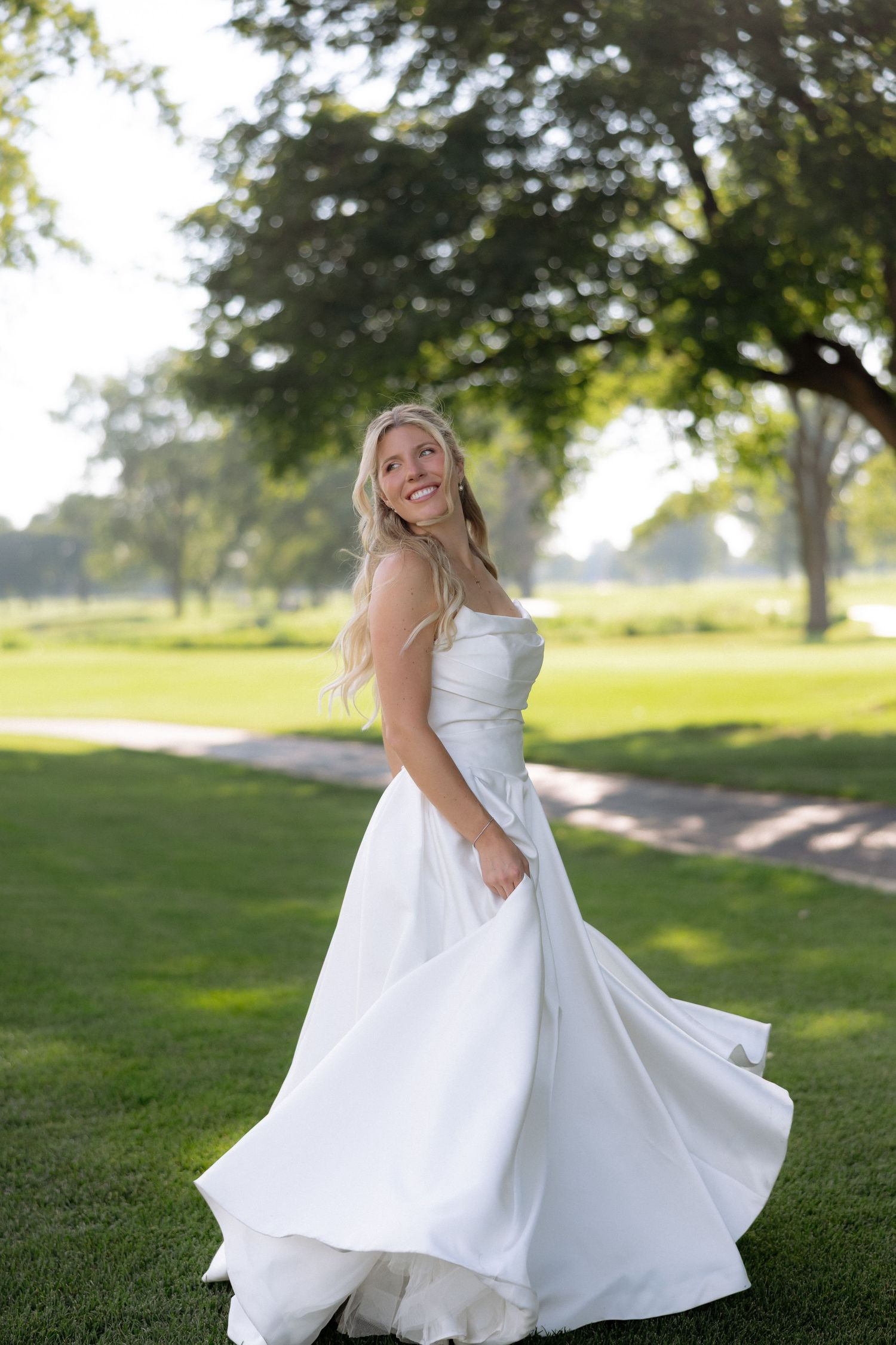 Smiling bride in strapless white wedding gown twirling in sunny outdoor park setting.