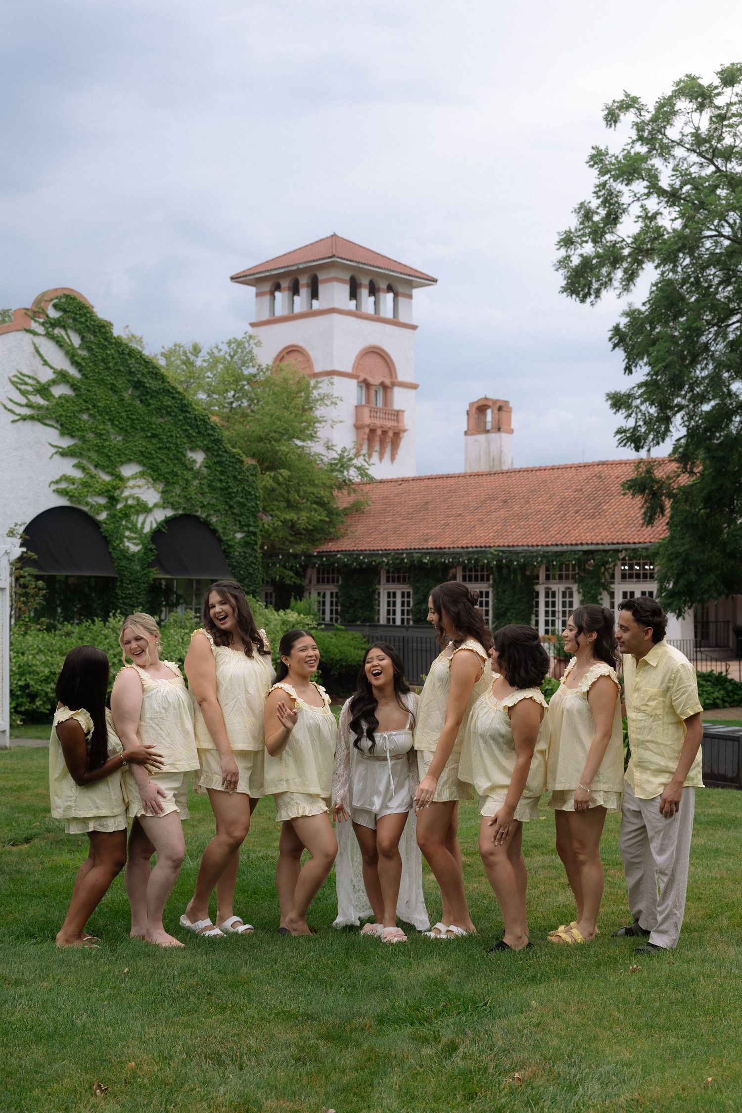 Bride in white surrounded by bridesmaids in yellow outfits laughing outdoors near a vine-covered building.