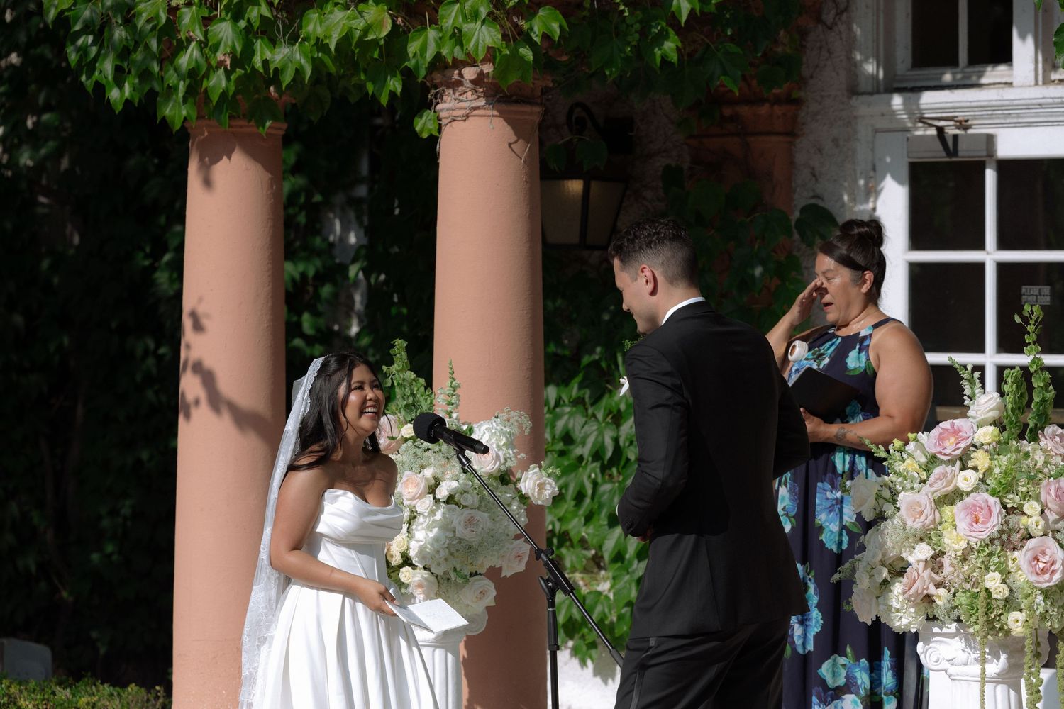Bride and groom exchange vows outdoors at elegant wedding ceremony with floral arrangements and officiant.
