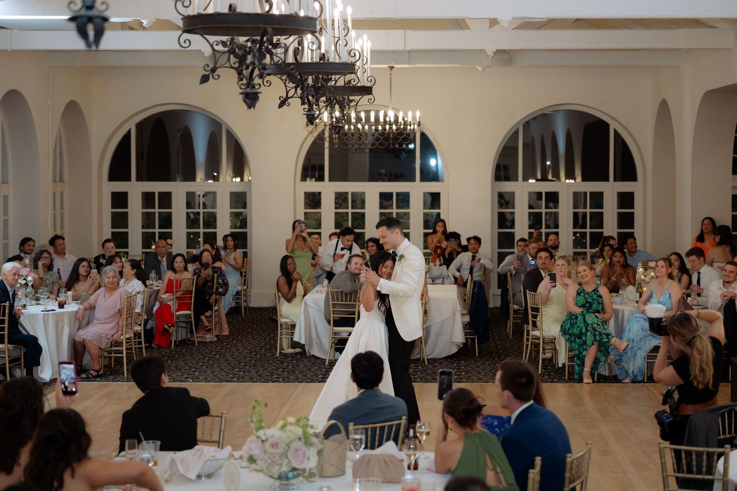Bride and groom share romantic first dance in elegant white ballroom while wedding guests watch from decorated tables.