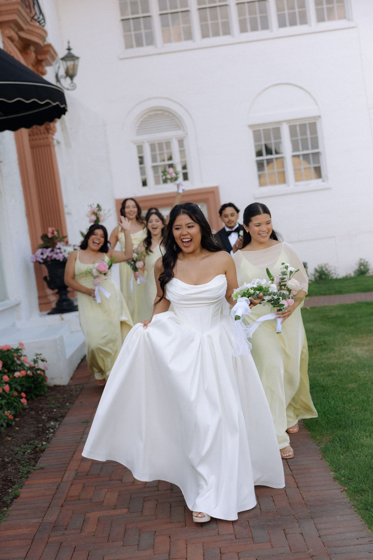 Joyful bride in strapless white gown lifting her dress while walking with bridesmaids outside a white venue.