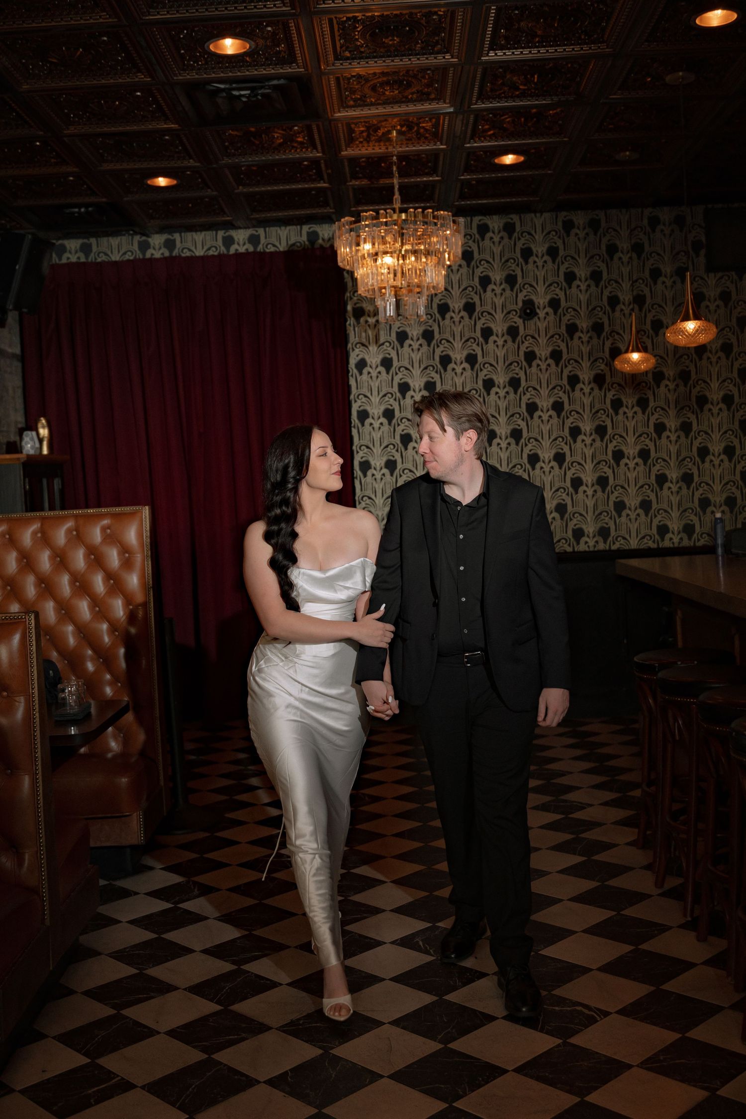Couple in elegant attire holding hands in a sophisticated bar with chandelier, checkered floors, and ornate wallpaper.
