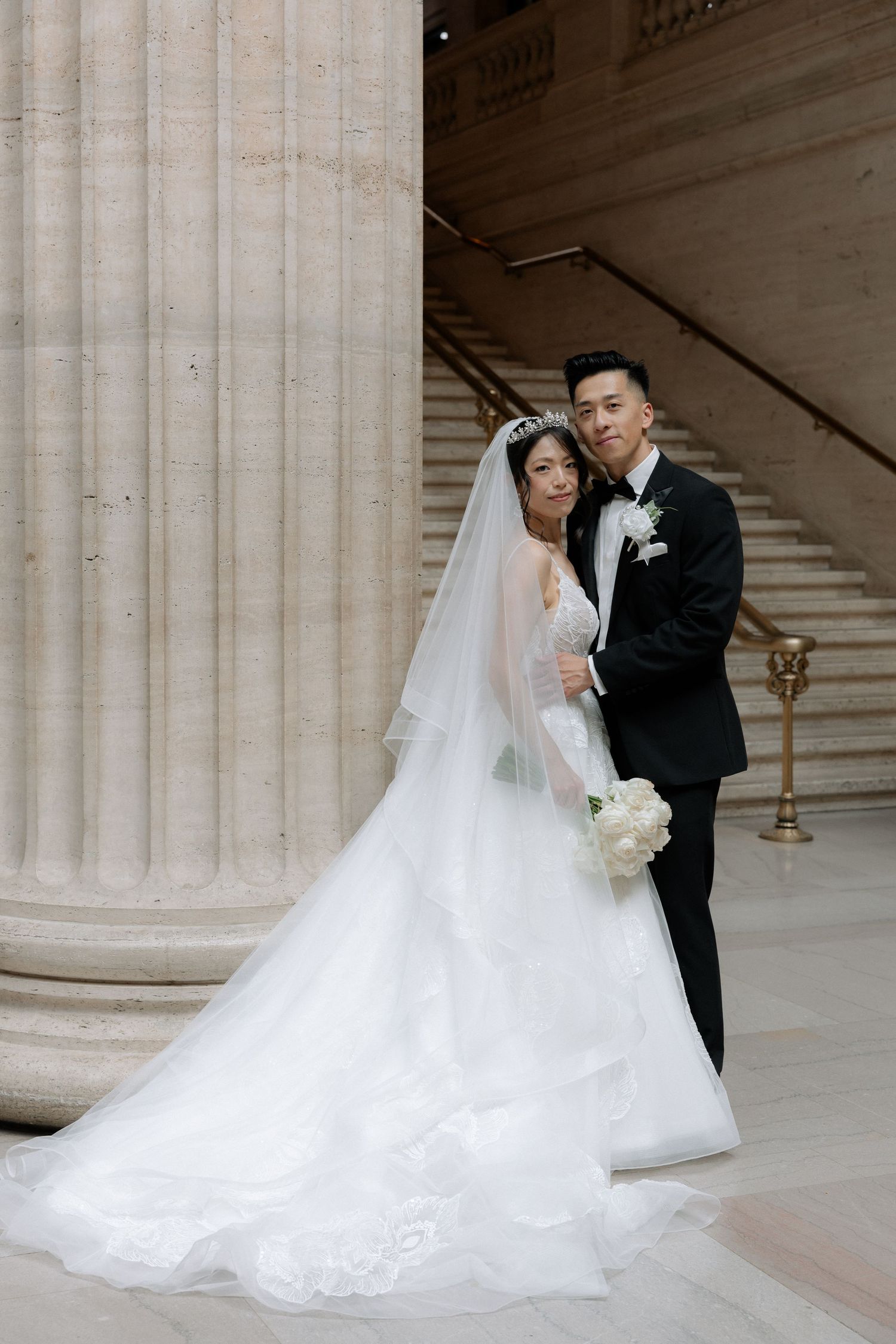 Asian bride in white ball gown with veil and groom in black tuxedo posing by marble columns and staircase.