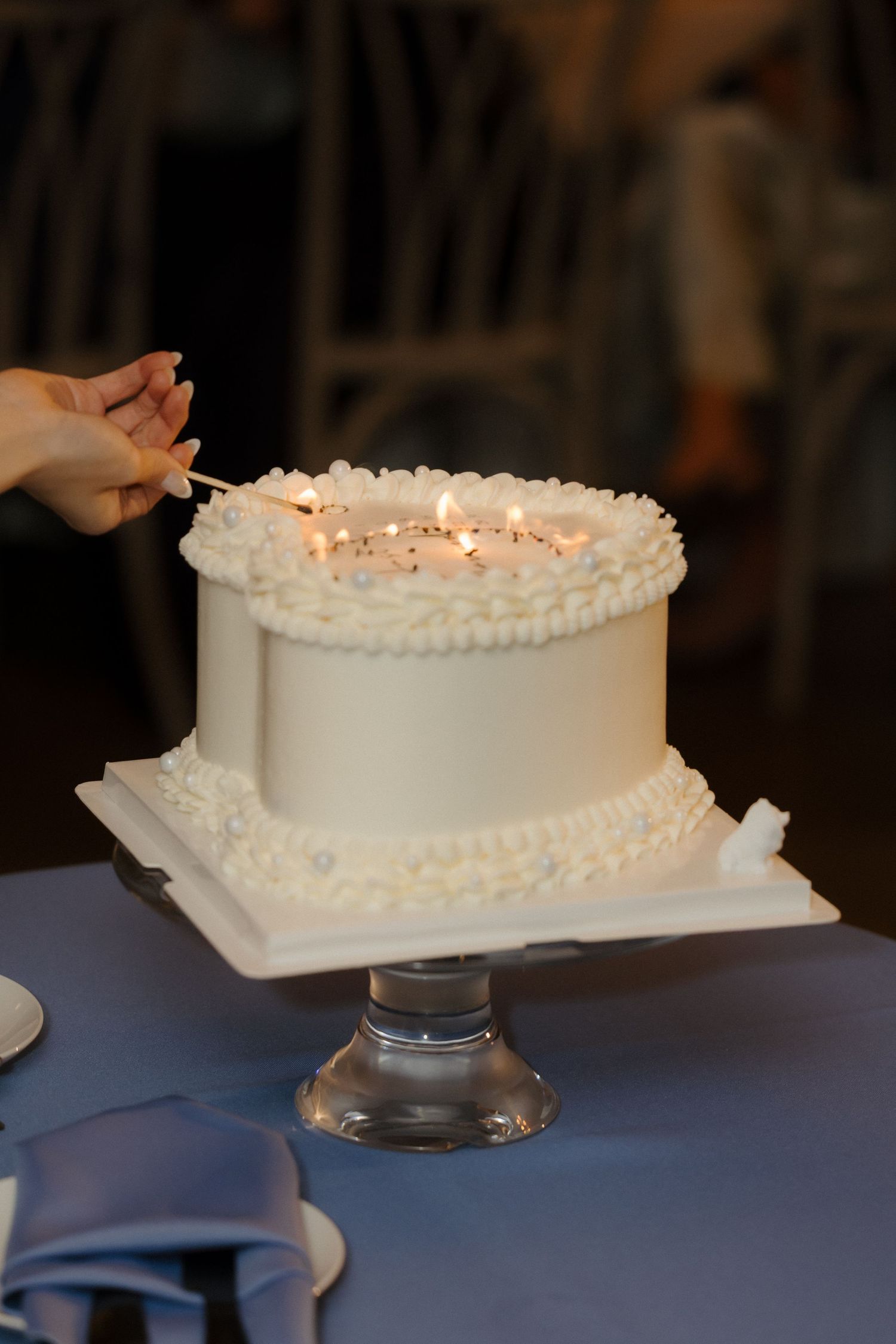 A white wedding cake with lit sparklers being ignited by hand on a glass pedestal stand.