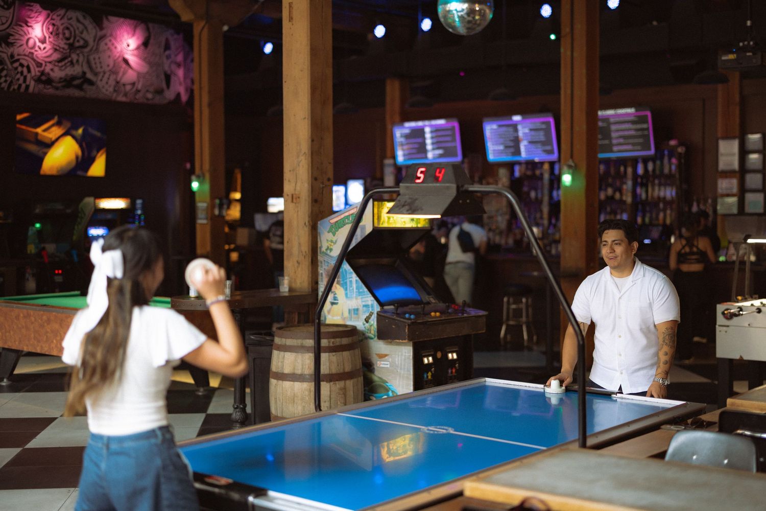 Couple playing air hockey at a retro arcade bar with colorful gaming machines in the background.