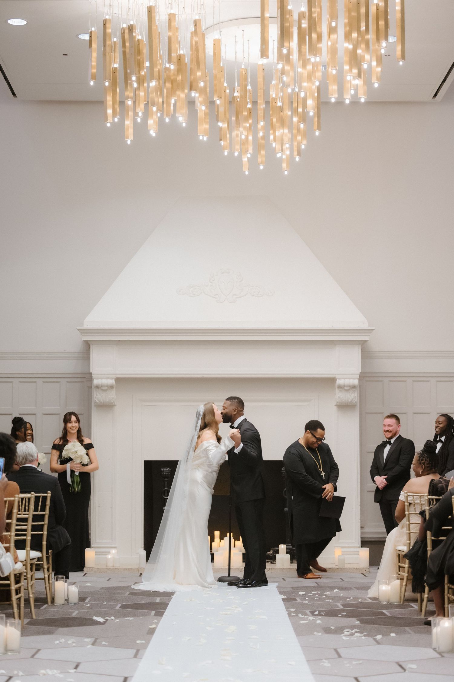 Bride and groom share first kiss at elegant indoor wedding ceremony with gold chandelier and white fireplace backdrop.