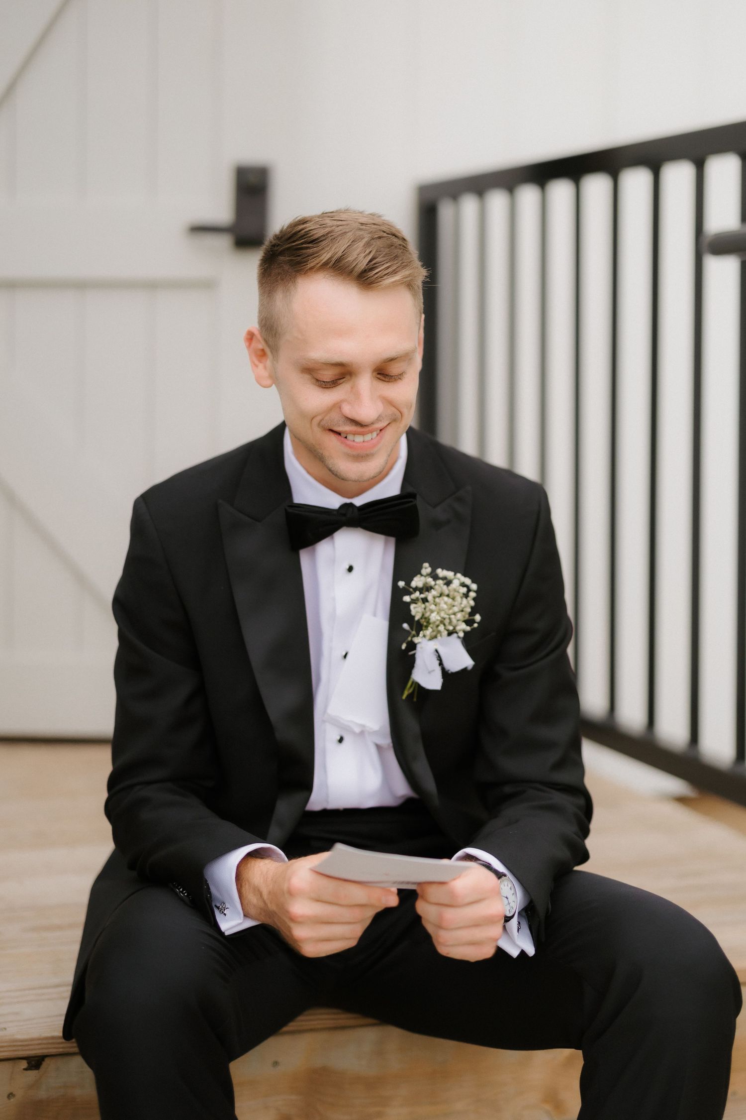 Happy groom in black tuxedo with boutonniere smiling while reading wedding day letter.