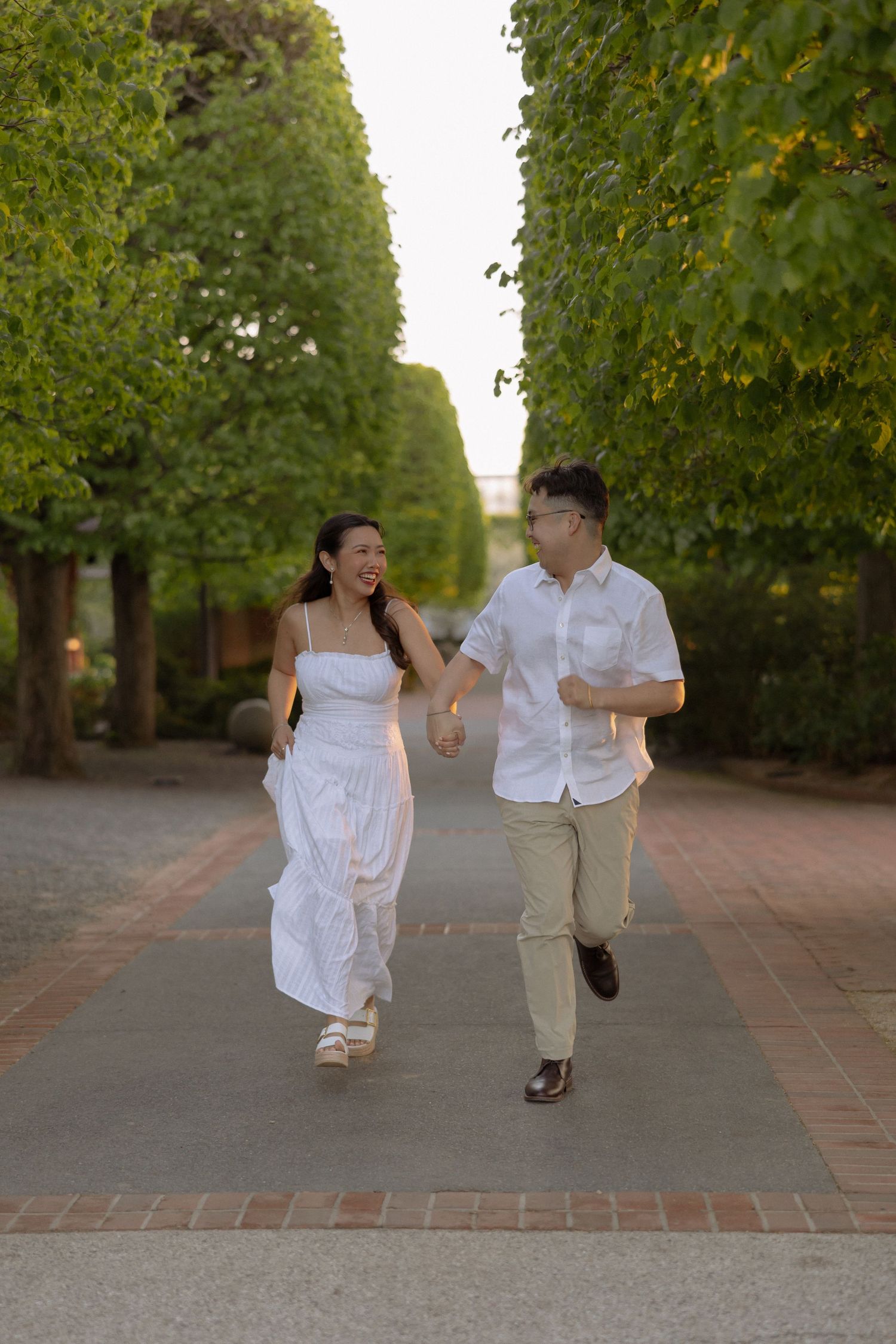 A joyful couple holds hands and runs along a tree-lined path, woman in white dress, man in white shirt and khakis.