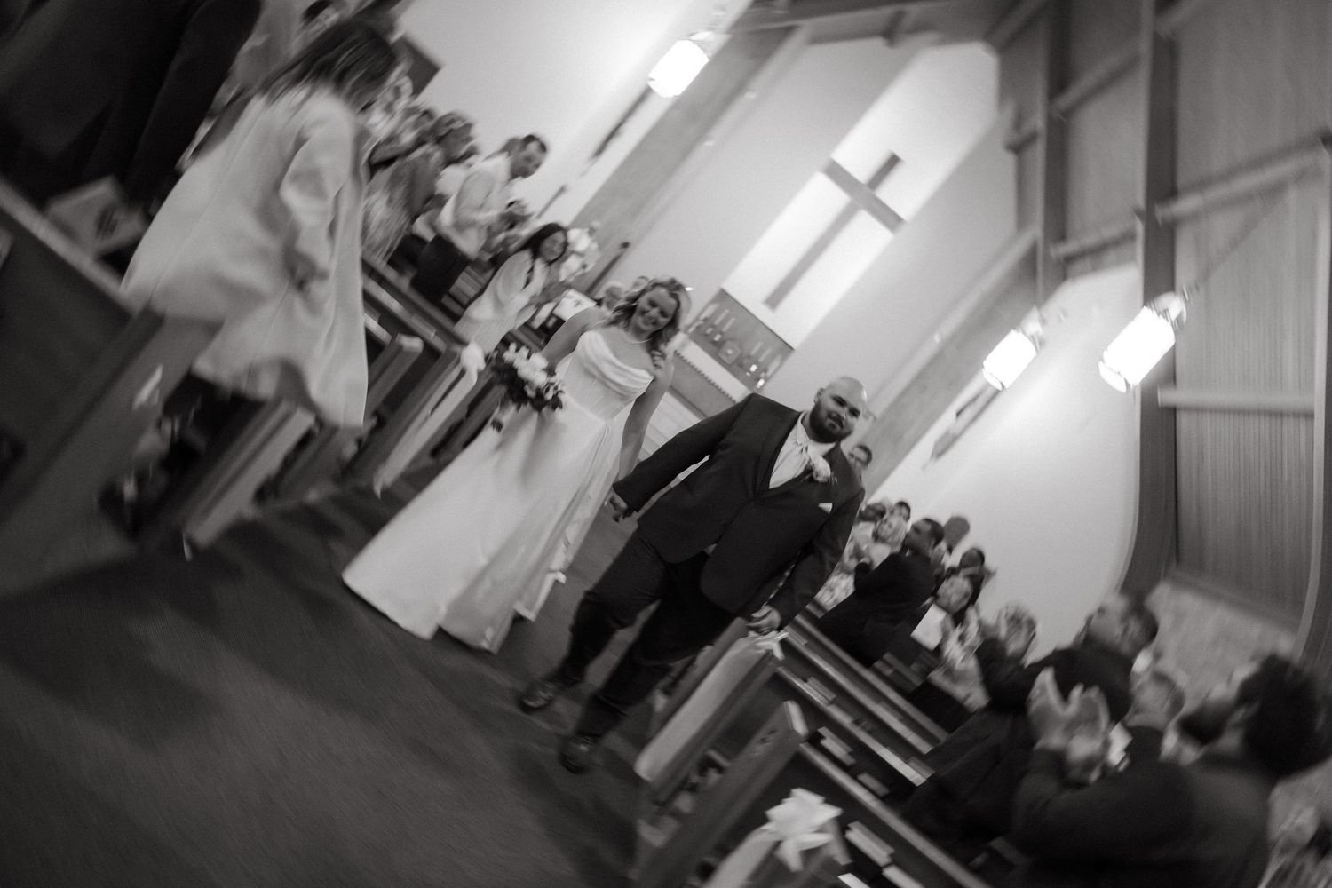 Black and white photo of a wedding ceremony inside a church with a cross on the wall and guests seated in pews.