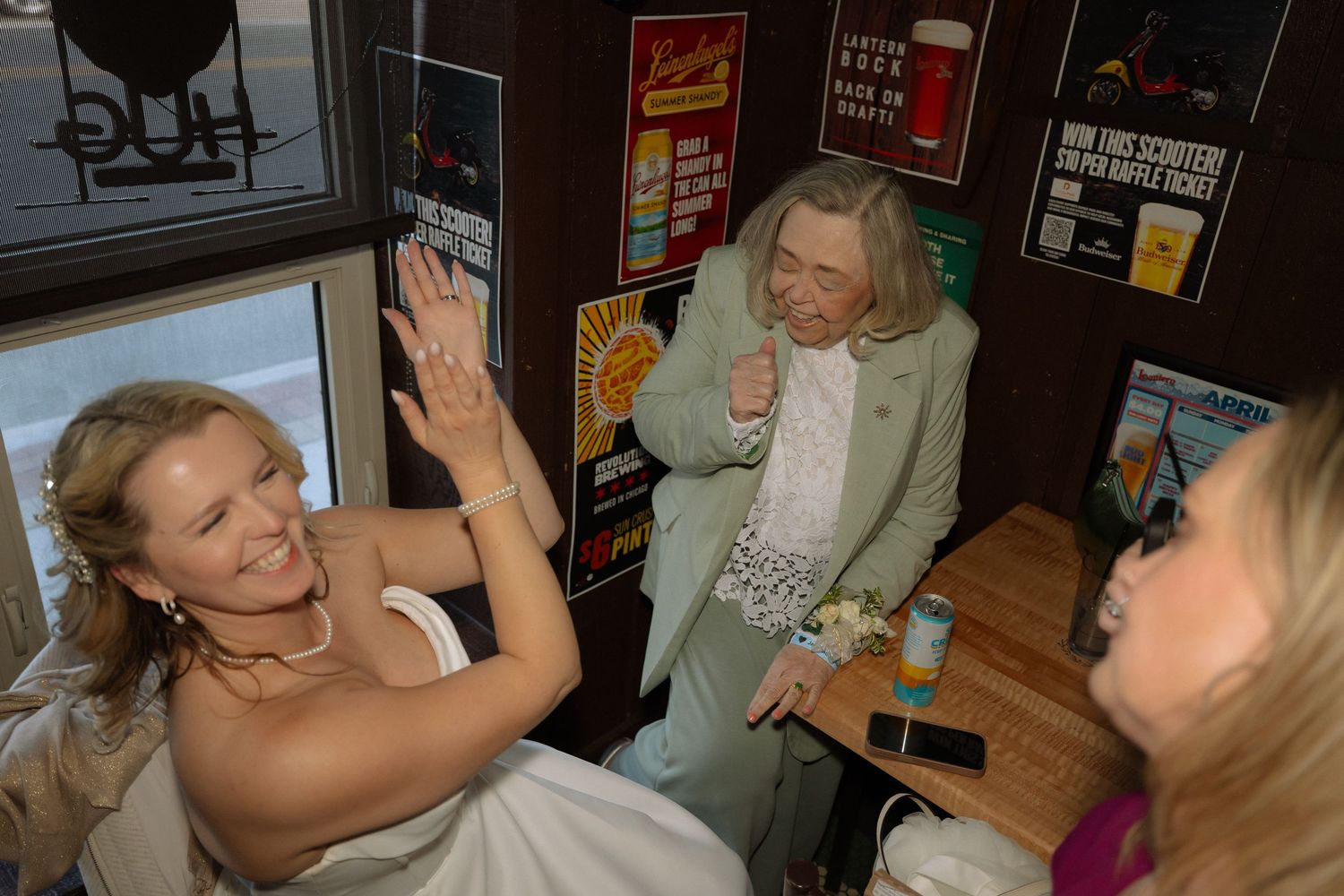 Bride in white dress laughing with guests at a bar decorated with colorful posters during a wedding celebration.