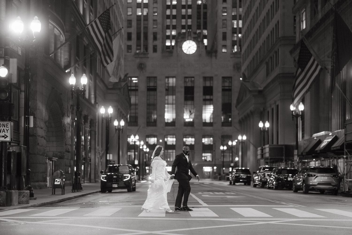 Black and white wedding photo of bride and groom crossing a city street at night with illuminated buildings.