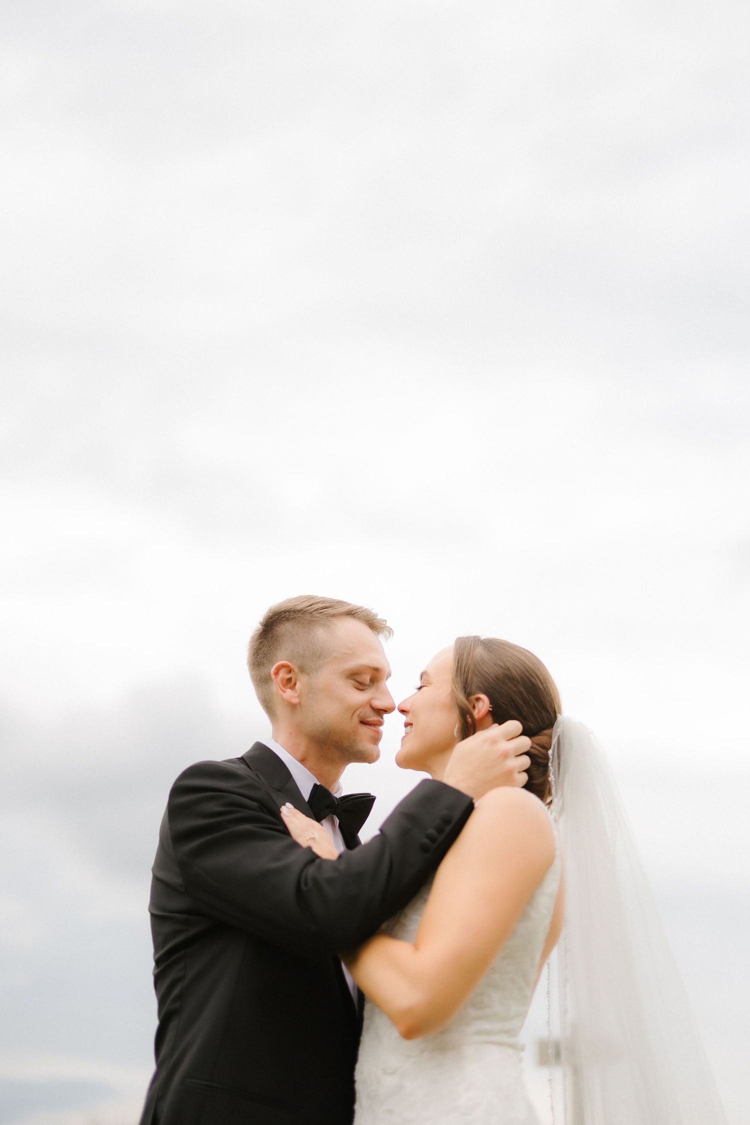 Bride and groom sharing an intimate almost-kiss moment under dramatic cloudy sky on wedding day.