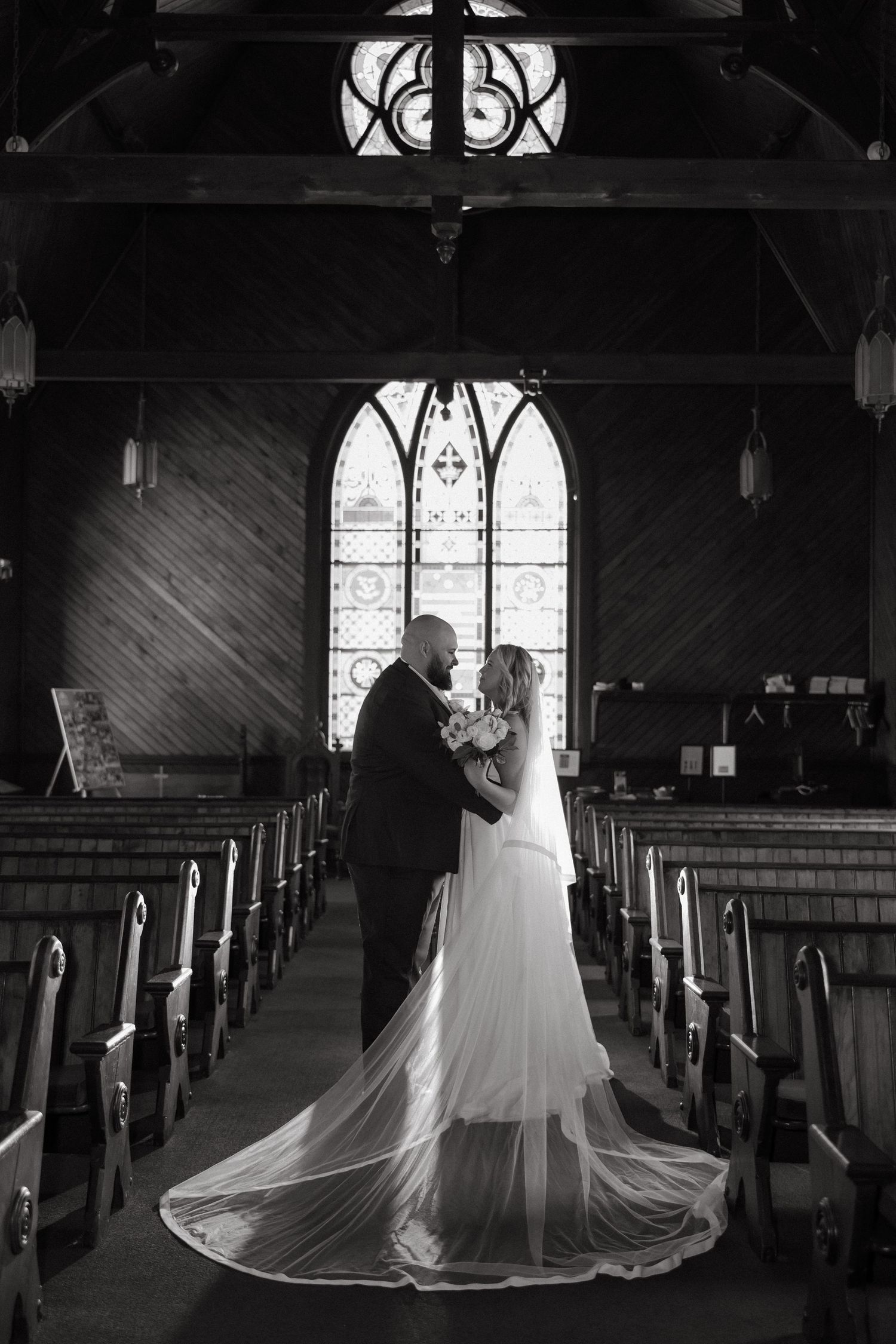 Black and white photo of bride and groom embracing at church altar with stained glass window and cathedral ceiling.