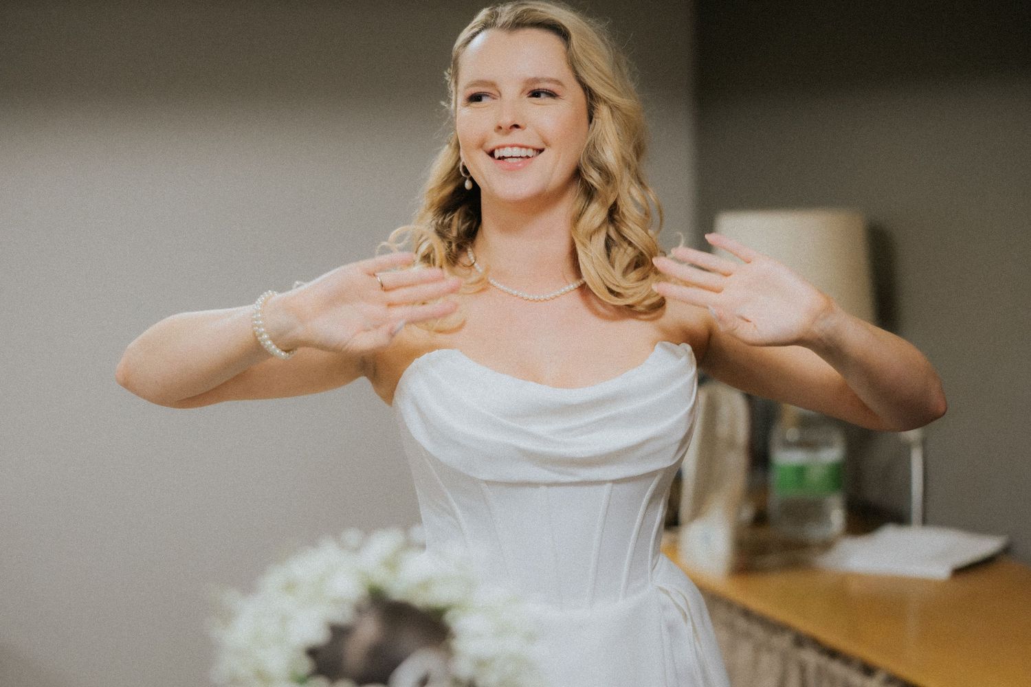 Smiling bride in white strapless wedding gown gesturing with hands, holding bouquet indoors.