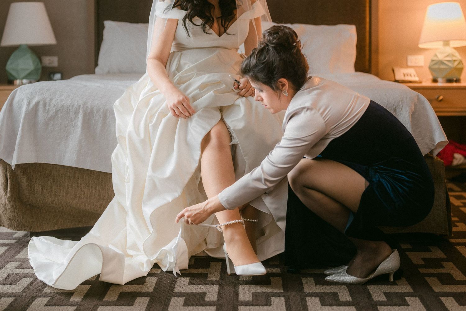 Bridesmaid kneels to help bride put on white heeled shoes while seated on a hotel bed.