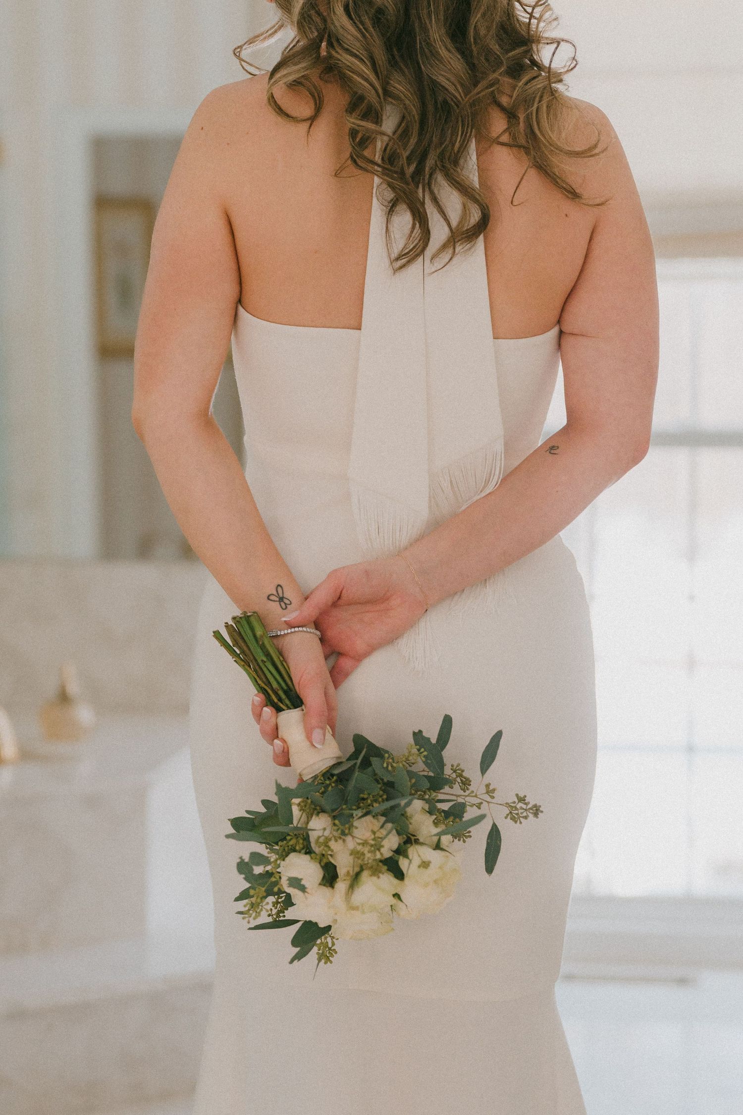 Bride in fitted white halter gown holding small white floral bouquet behind her back.