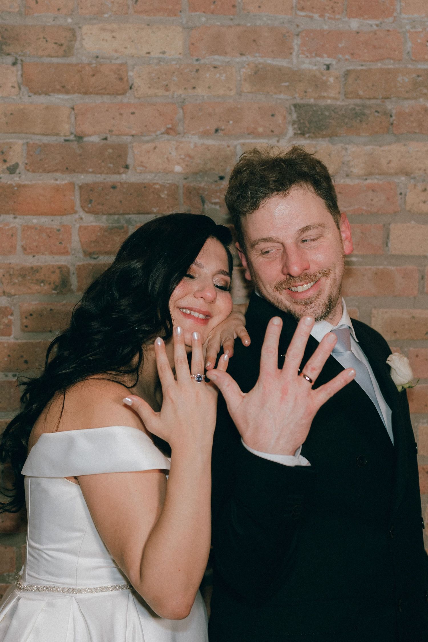 Newlywed couple smiling and showing off their wedding rings against a rustic brick wall backdrop.