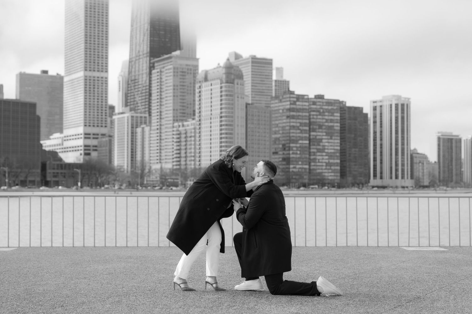 Black and white photo of couple embracing after marriage proposal on Chicago lakefront with skyline.