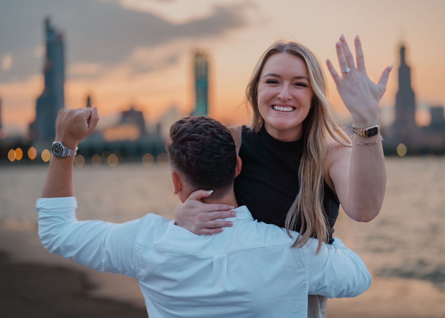 Couple celebrating engagement at sunset with city skyline, woman showing ring while being lifted by partner near waterfront.