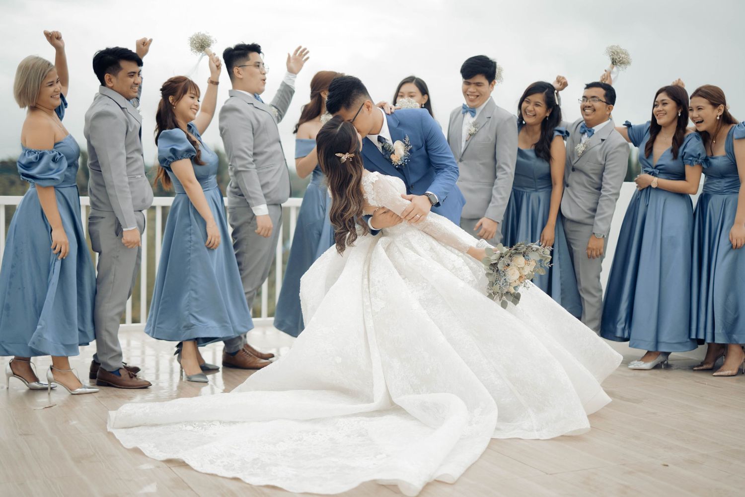 A couple shares a romantic kiss in wedding attire surrounded by bridesmaids in blue dresses and groomsmen in gray suits.