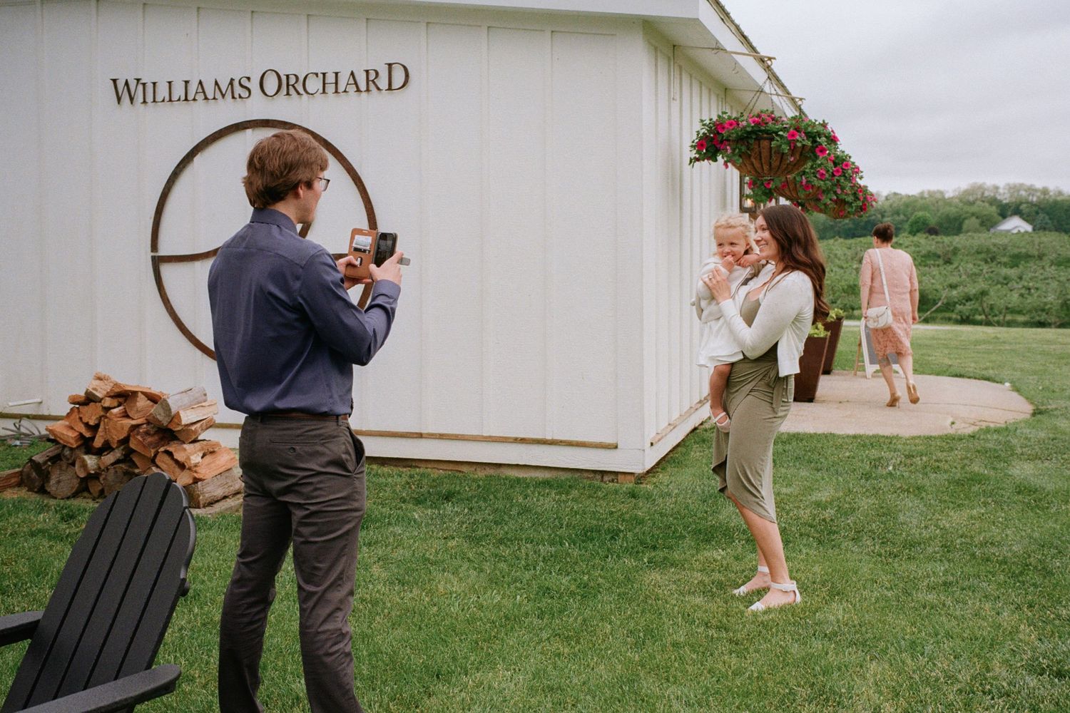 A man photographs a woman holding a baby outside Williams Orchard white barn venue on a cloudy day.