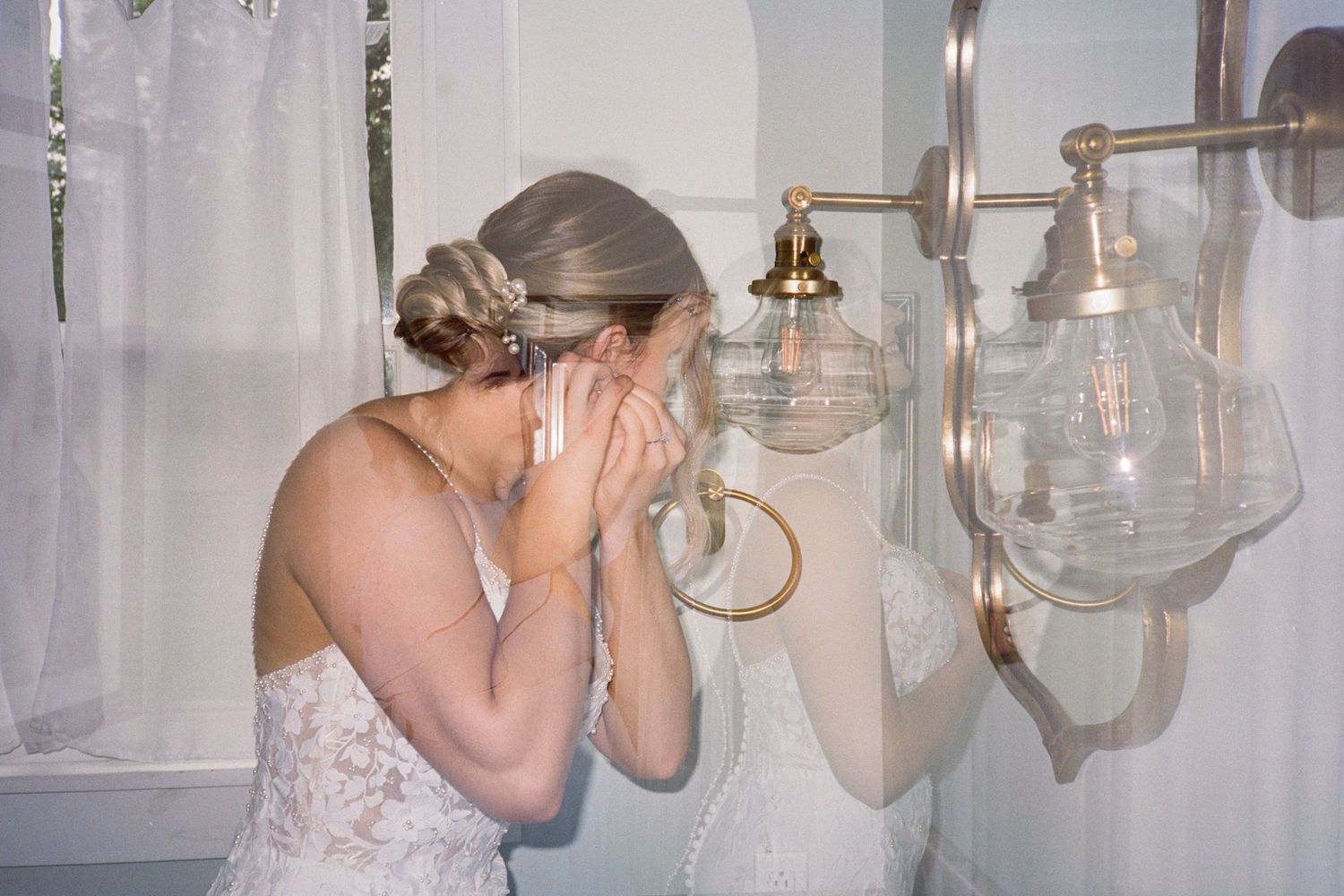Emotional bride in lace wedding dress covering her face near gold wall sconces and mirror in elegant bathroom.