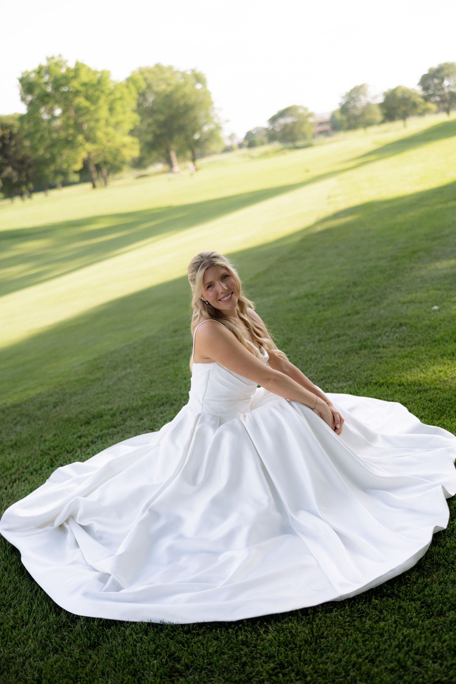 Smiling bride in strapless white ball gown sitting on lush green grass at outdoor venue.