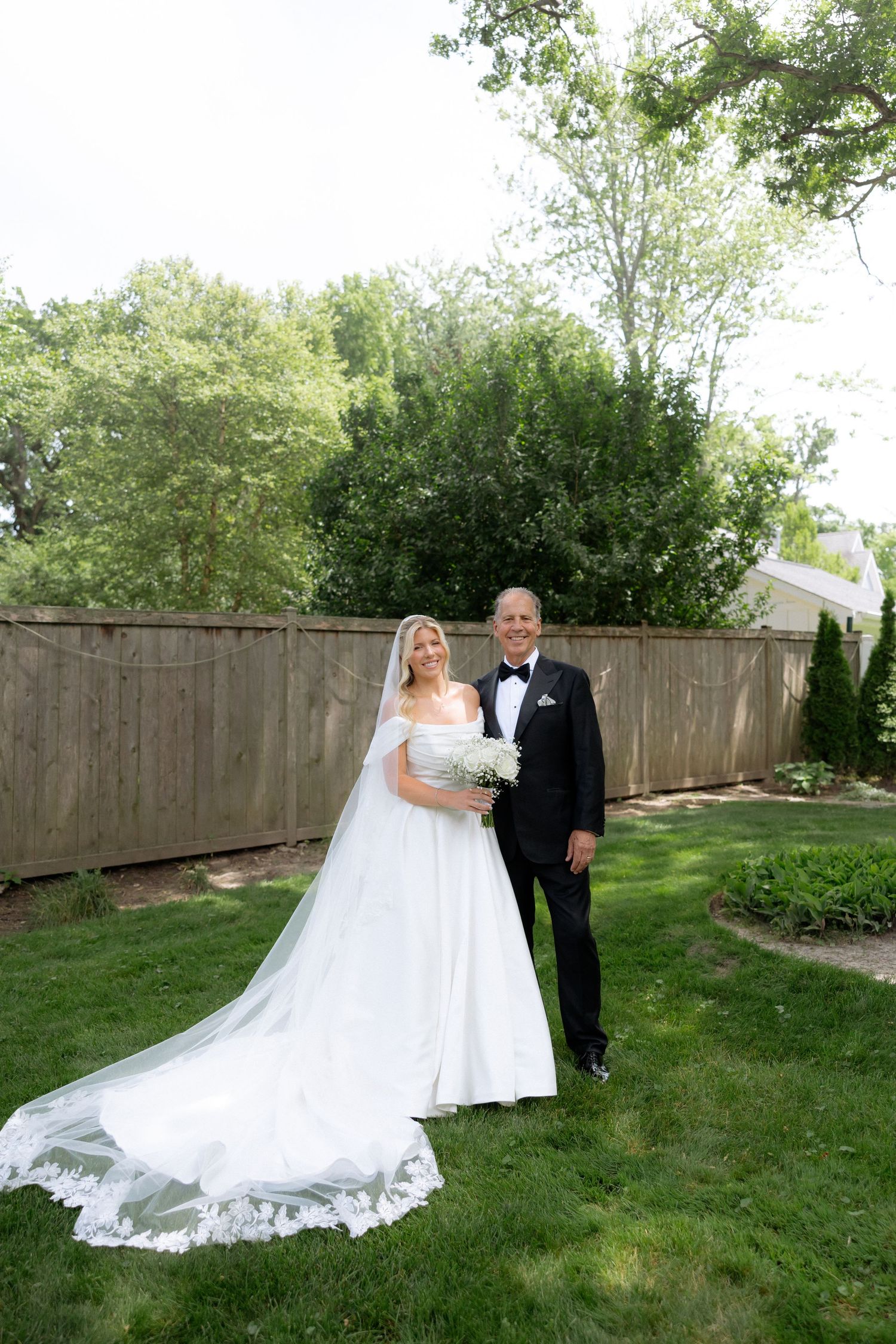 Bride in flowing white gown with cathedral veil poses with father in tuxedo in garden.