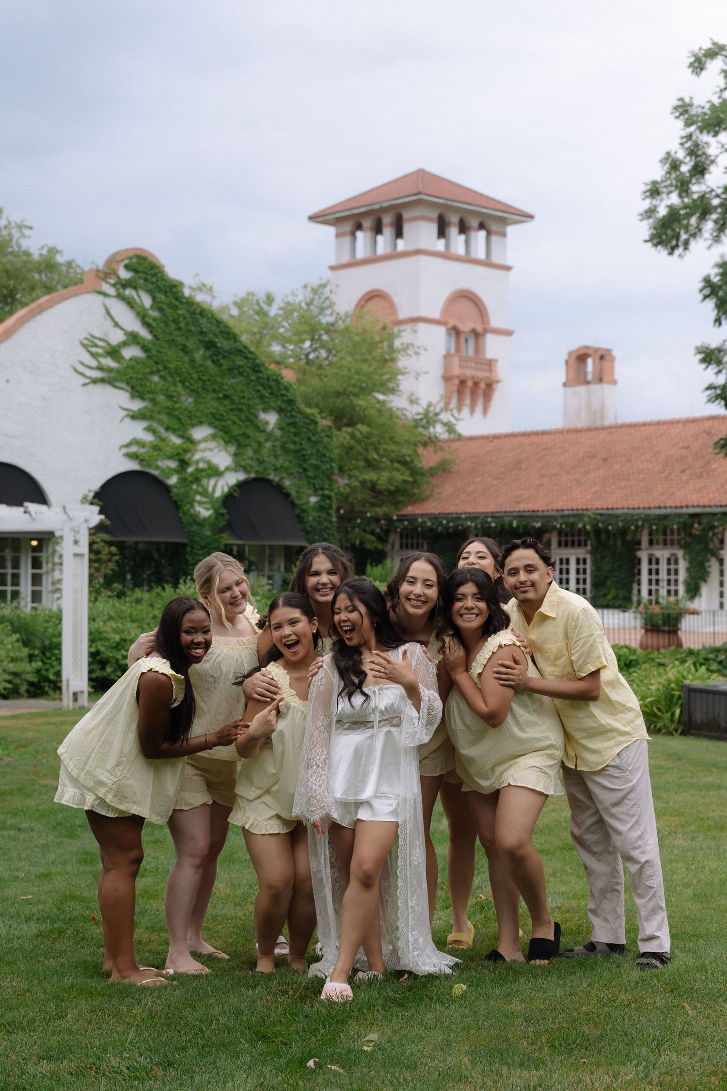 Bride in white robe poses with bridesmaids in yellow outfits on green lawn outside elegant white venue.