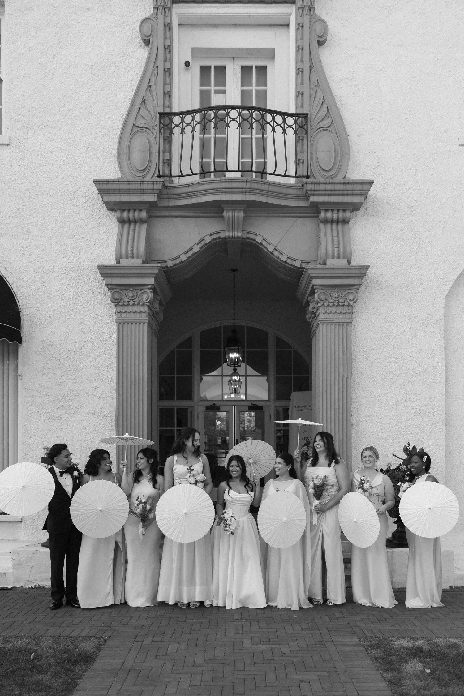 Bridal party in white gowns holding parasols pose outside elegant venue entrance in black and white photo.