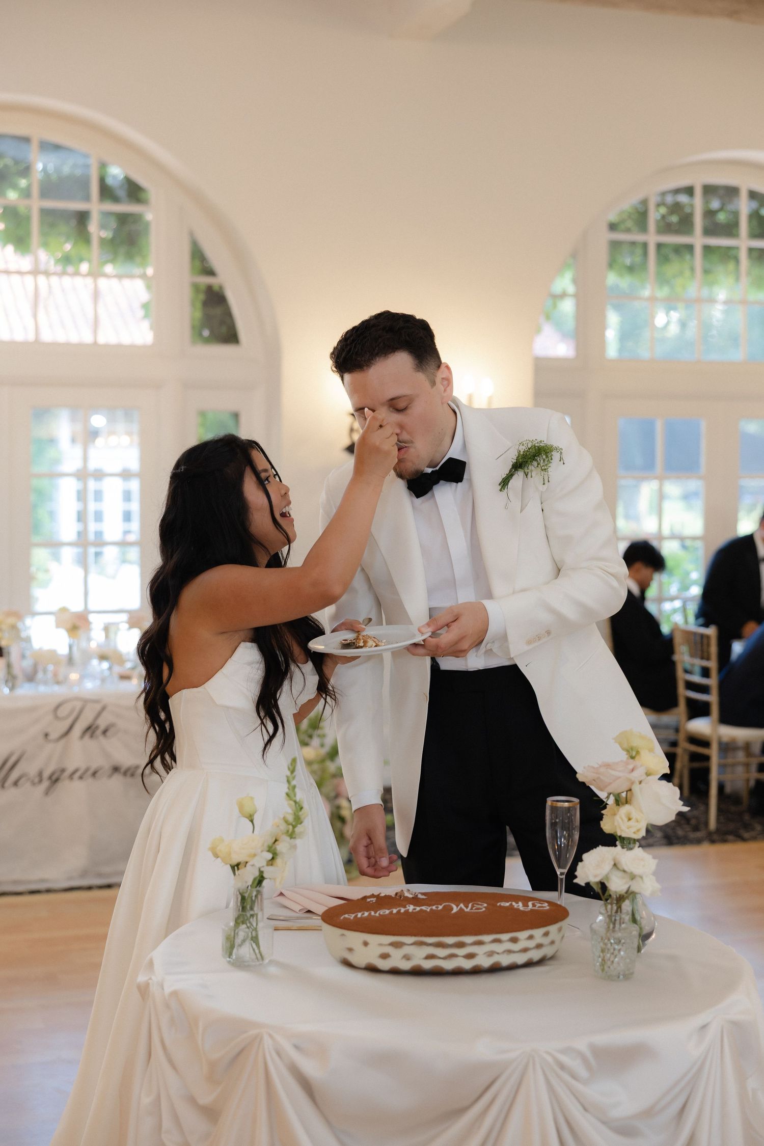 Bride feeding groom wedding cake at reception sweetheart table in elegant white ballroom venue.