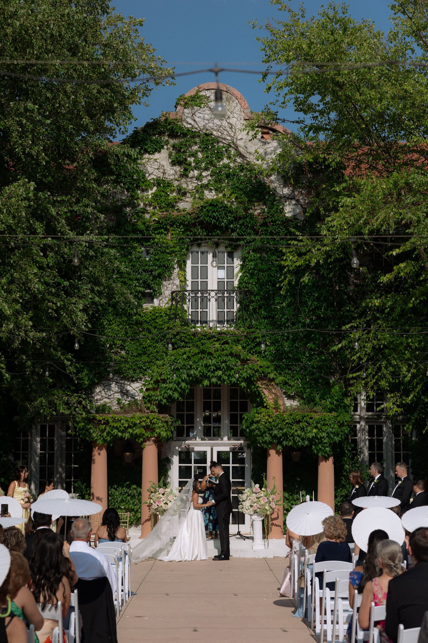 Outdoor wedding ceremony at ivy-covered historic venue with guests seated in white chairs under blue sky.