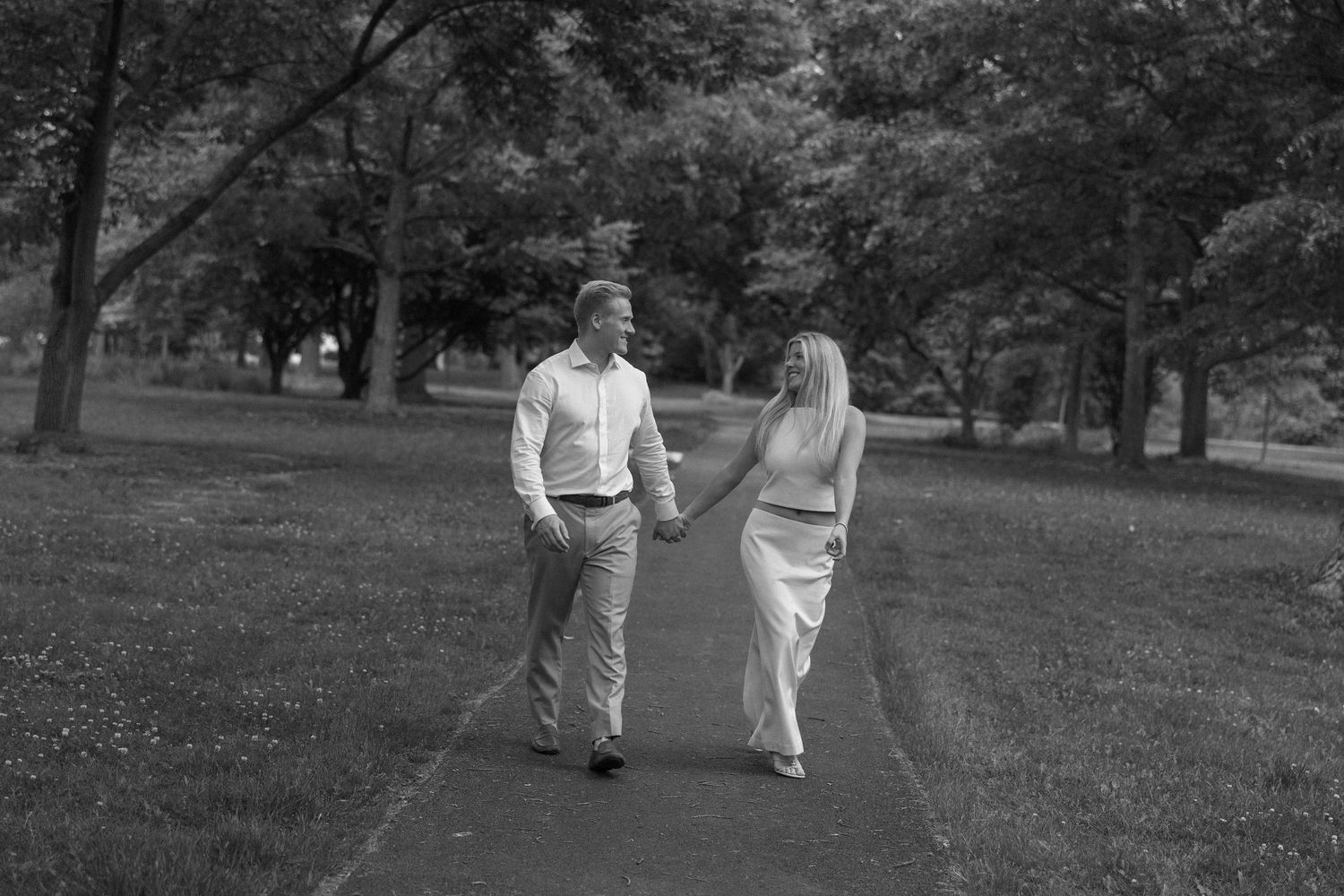 Black and white photo of a couple holding hands and walking on a path through a lush green park.